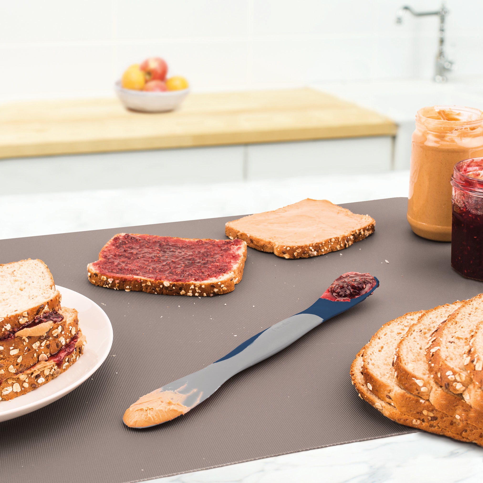 On a kitchen counter, slices of bread—one with jelly, one with peanut butter—sit next to peanut butter and jelly jars, stacked sandwiches, and the Scoop and Spread tool, perfect for scooping and spreading both.