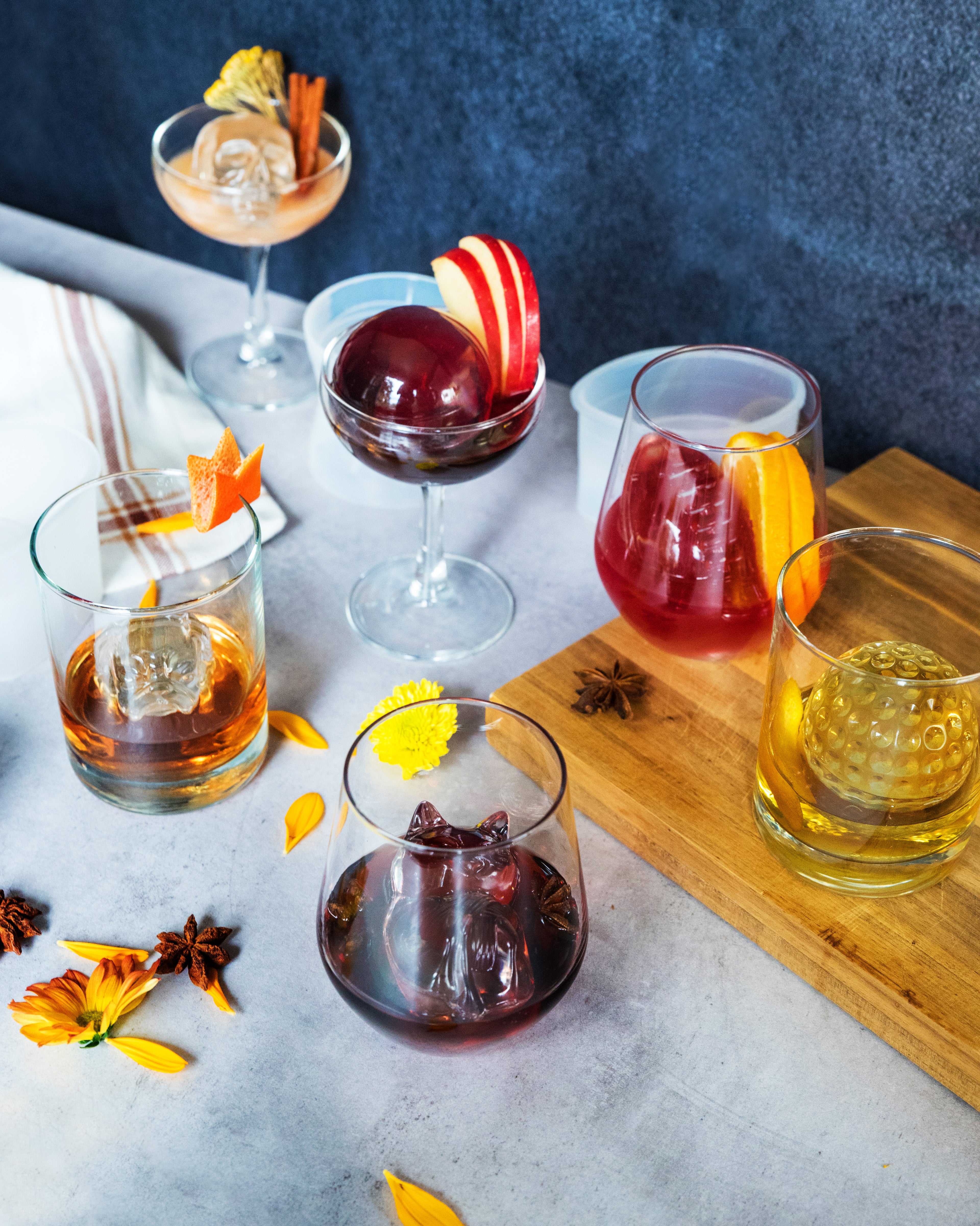 A variety of vibrant cocktails in assorted glasses, each topped with fruits or spices and cooled using Designer Ice Mold shapes, are arranged on a table decorated with flower petals and star anise.