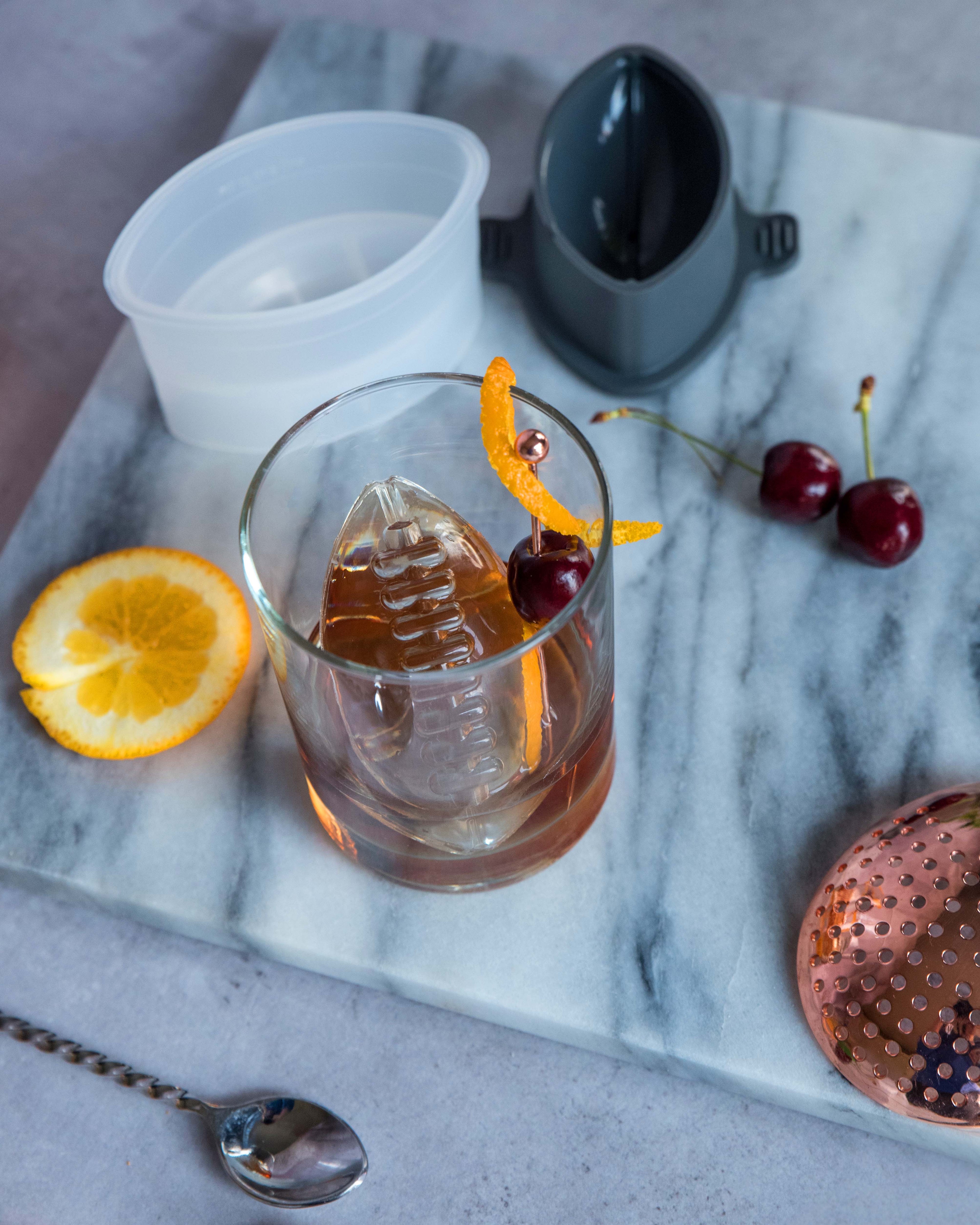 A cocktail in a glass with a large clear ice shape made using the Sports Ice Mold, garnished with orange peel and cherry, sits on a marble board surrounded by sliced orange, cherries, the Sports Ice Mold, a strainer, and a cocktail spoon.