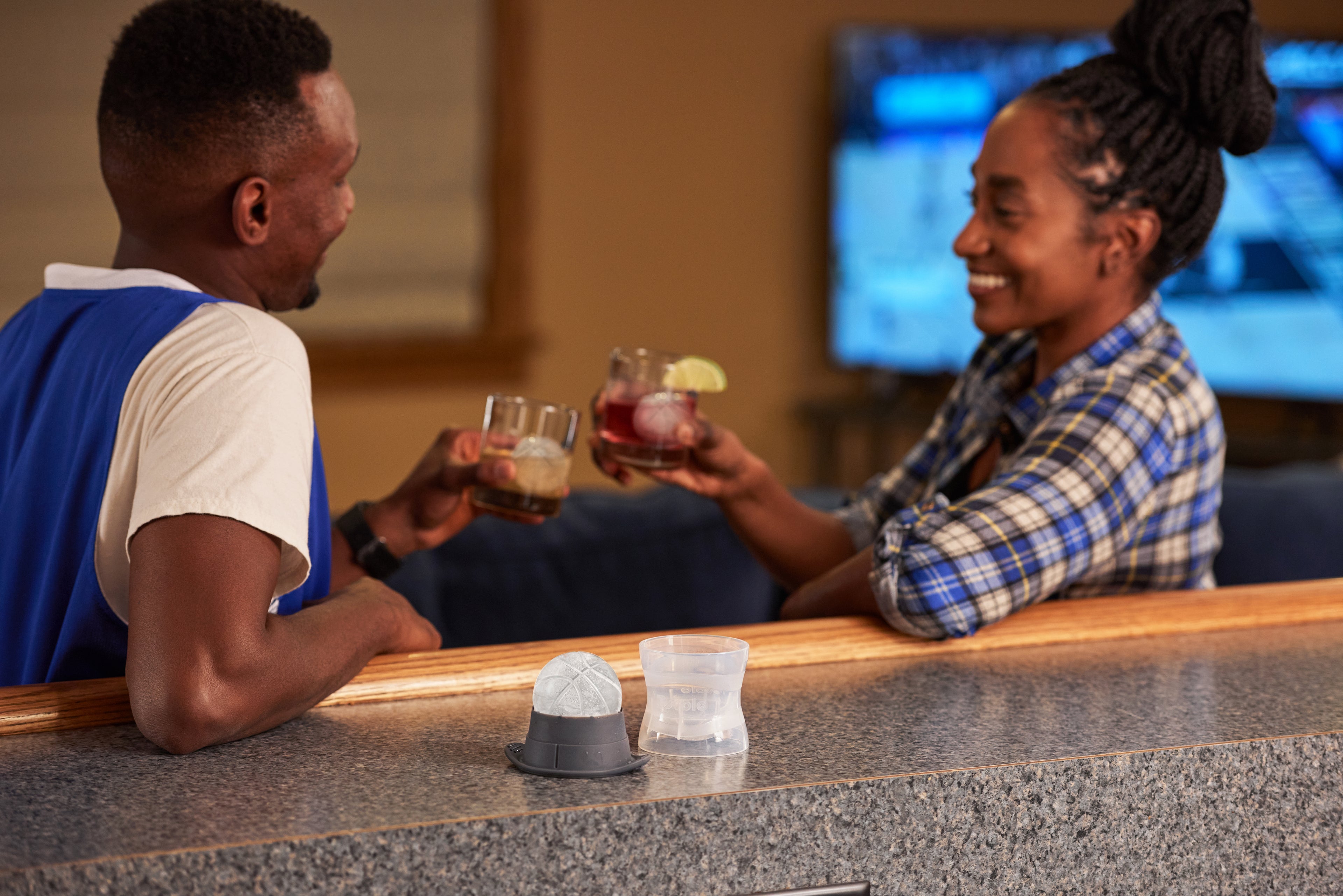 Two people smile and clink glasses with Sports Ice Mold creations while sitting at a counter in a cozy home. A TV plays in the background, and a small sensor device is visible on the counter in the foreground.