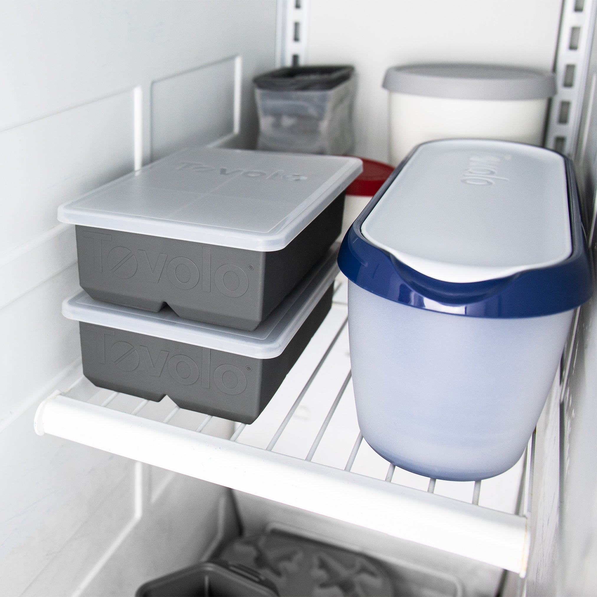 A King Cube Ice Mold, along with two stacked gray trays and a blue-and-white lidded container, are arranged on a white wire freezer shelf with additional storage items in the background.