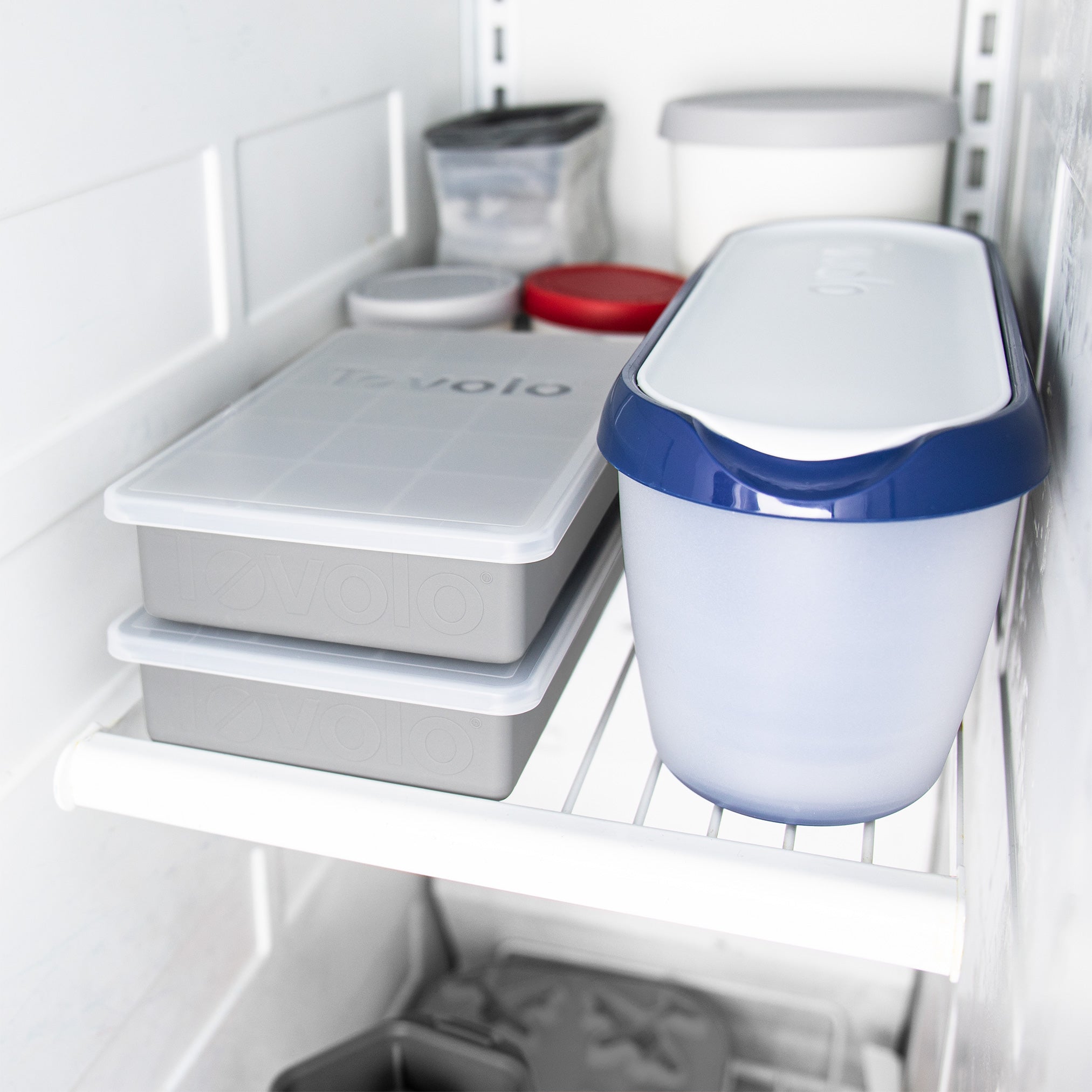 The Perfect Cube Ice Mold is stored on a wire shelf inside the freezer, alongside various plastic containers with lids, ice cube trays, and a silicone tray for making long-lasting ice in different shapes and sizes.