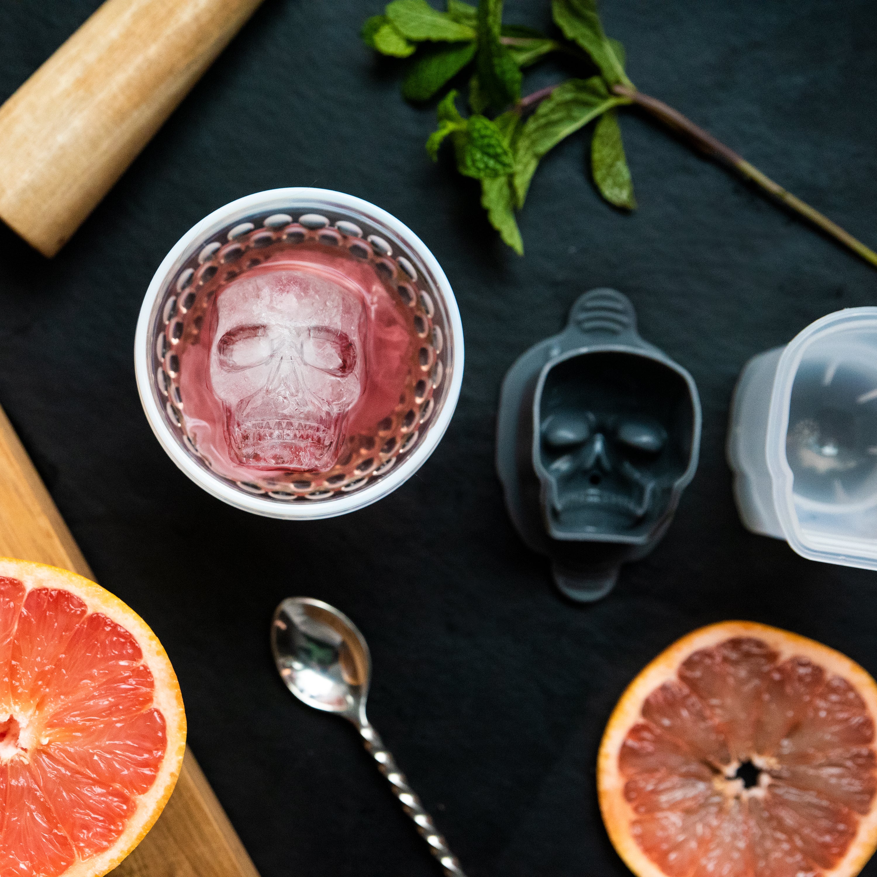 A drink with a skull-shaped ice cube, made using the Designer Ice Mold, sits on a dark surface surrounded by grapefruit, mint leaves, a spoon, a muddler, and an open Designer Ice Mold.