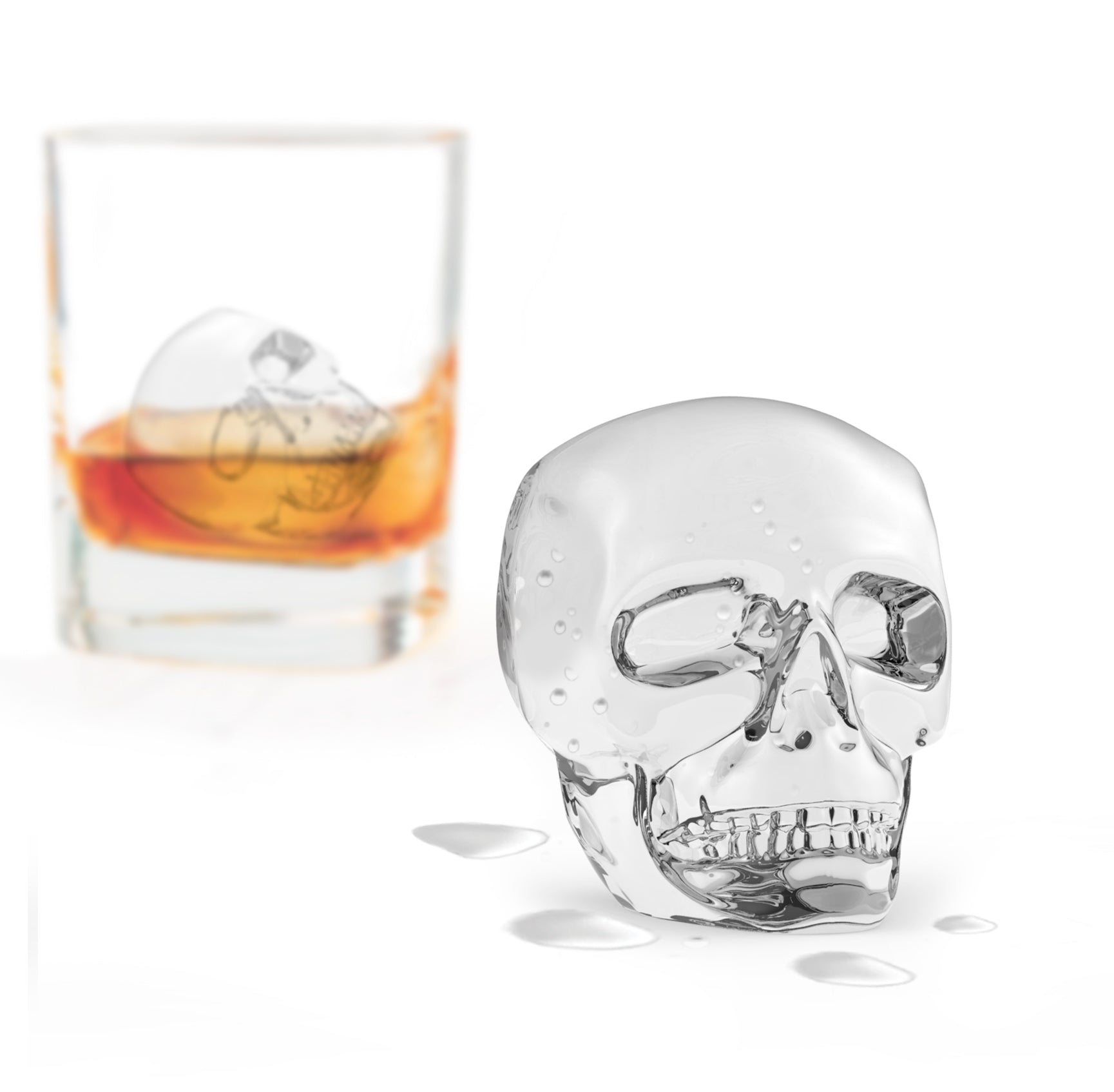 A large skull-shaped ice cube made with the Designer Ice Mold melts on a white surface, water droplets around it. In the background, a glass of amber liquid holds another skull-shaped ice cube.