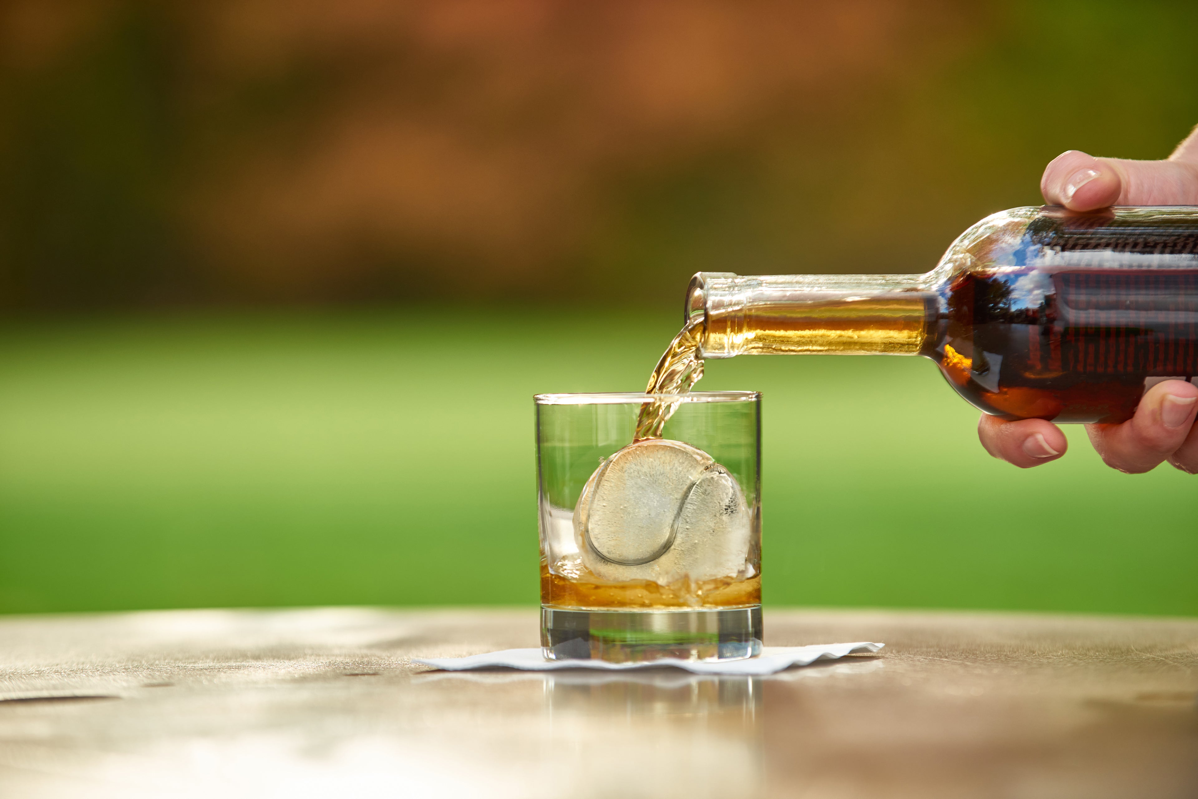 A hand pours whiskey into a glass with a large round ice cube made from the Sports Ice Mold, set on a napkin outdoors against a blurred green background.