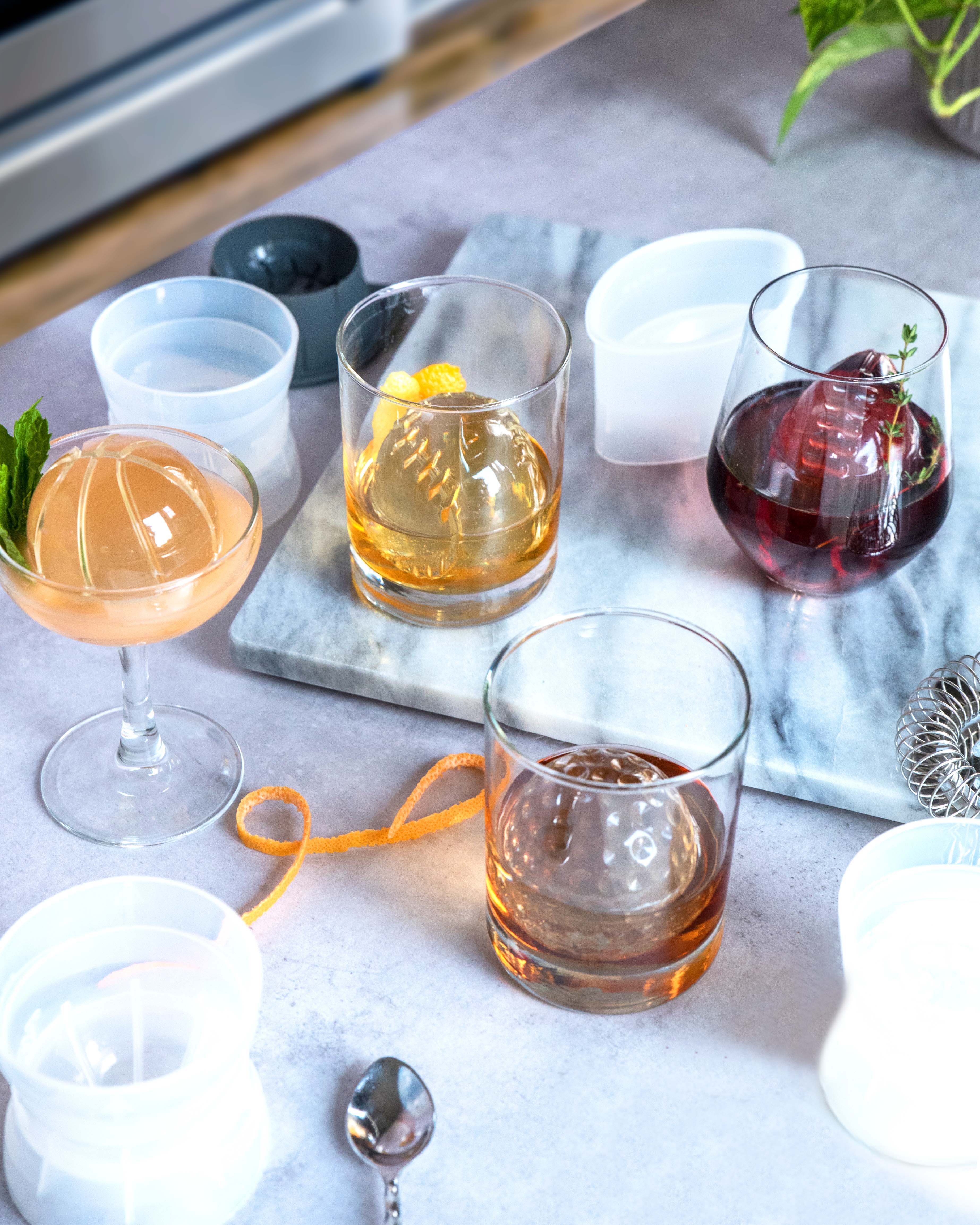 Four cocktails with large, clear ice spheres made with the Sports Ice Mold are arranged on a marble tray, accompanied by the molds, a cocktail strainer, spoon, and orange peel garnish atop a light countertop.