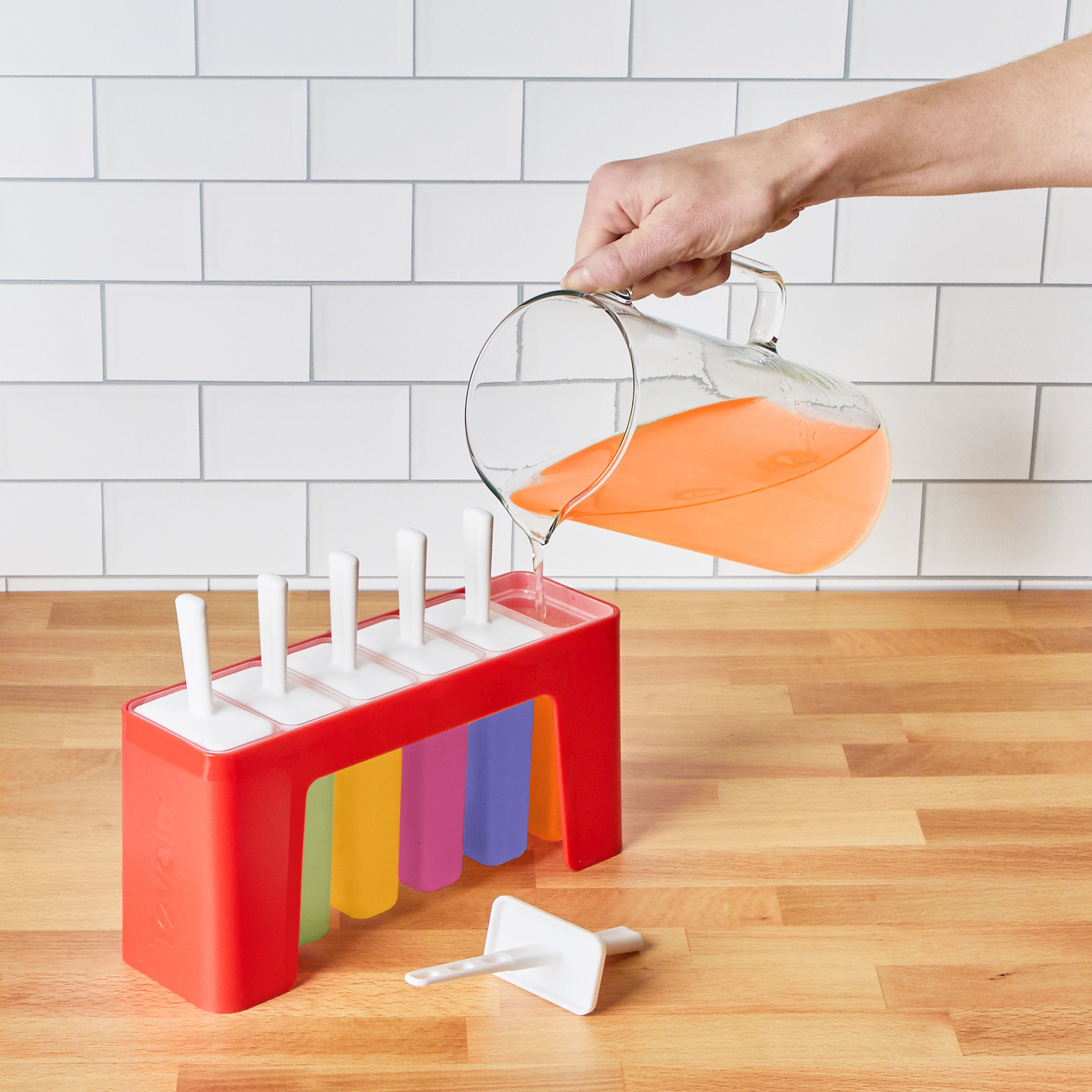 A hand pours orange liquid from a glass pitcher into Pop Molds w/ Tray, featuring colorful containers on a wooden counter with one lid and white stick in front—ideal for making homemade frozen treats.