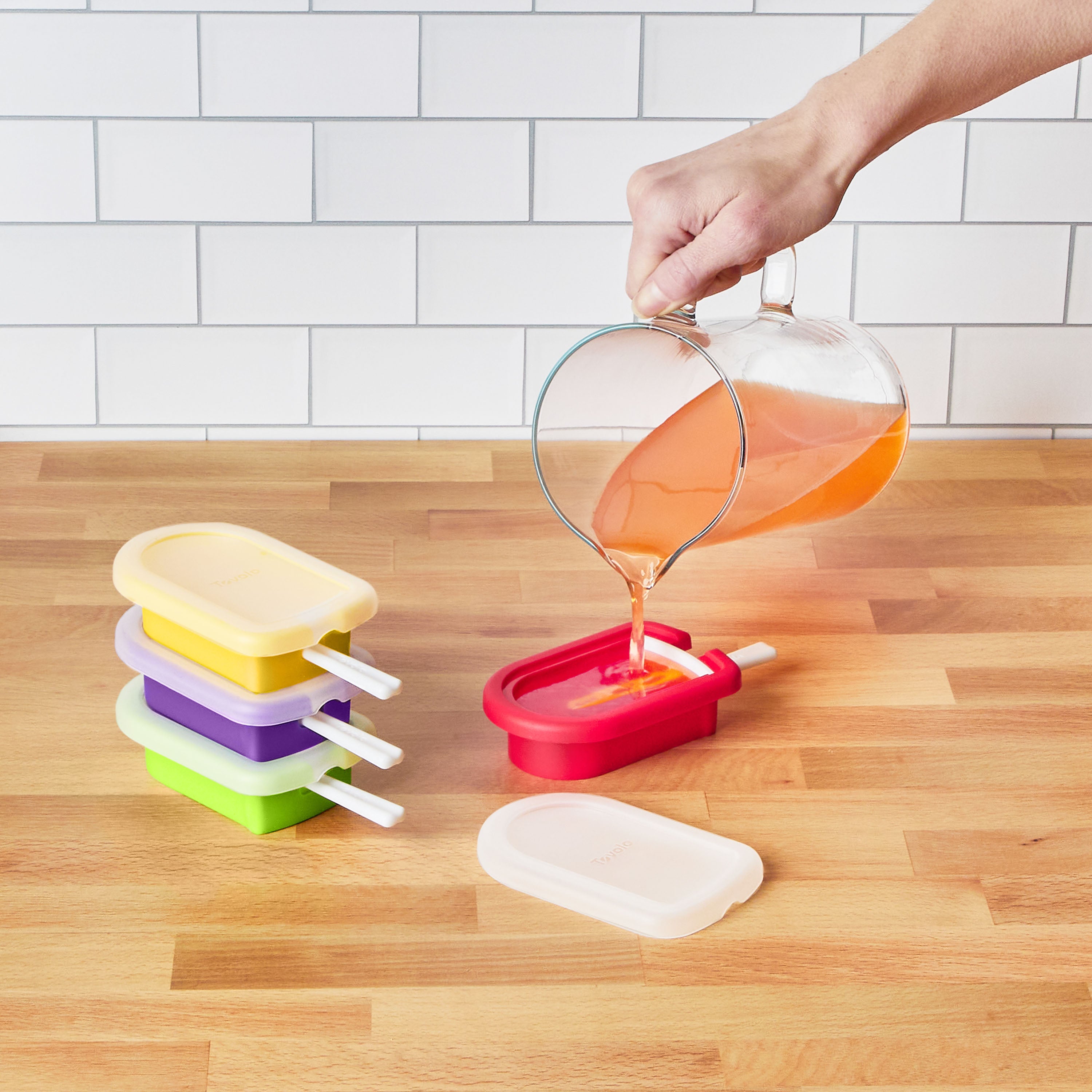 A hand pours orange liquid from a glass measuring cup into a red Pop Molds Stackable popsicle maker on a wooden countertop, with three colorful stacked Pop Molds Stackable makers and a white tile backsplash in the background.