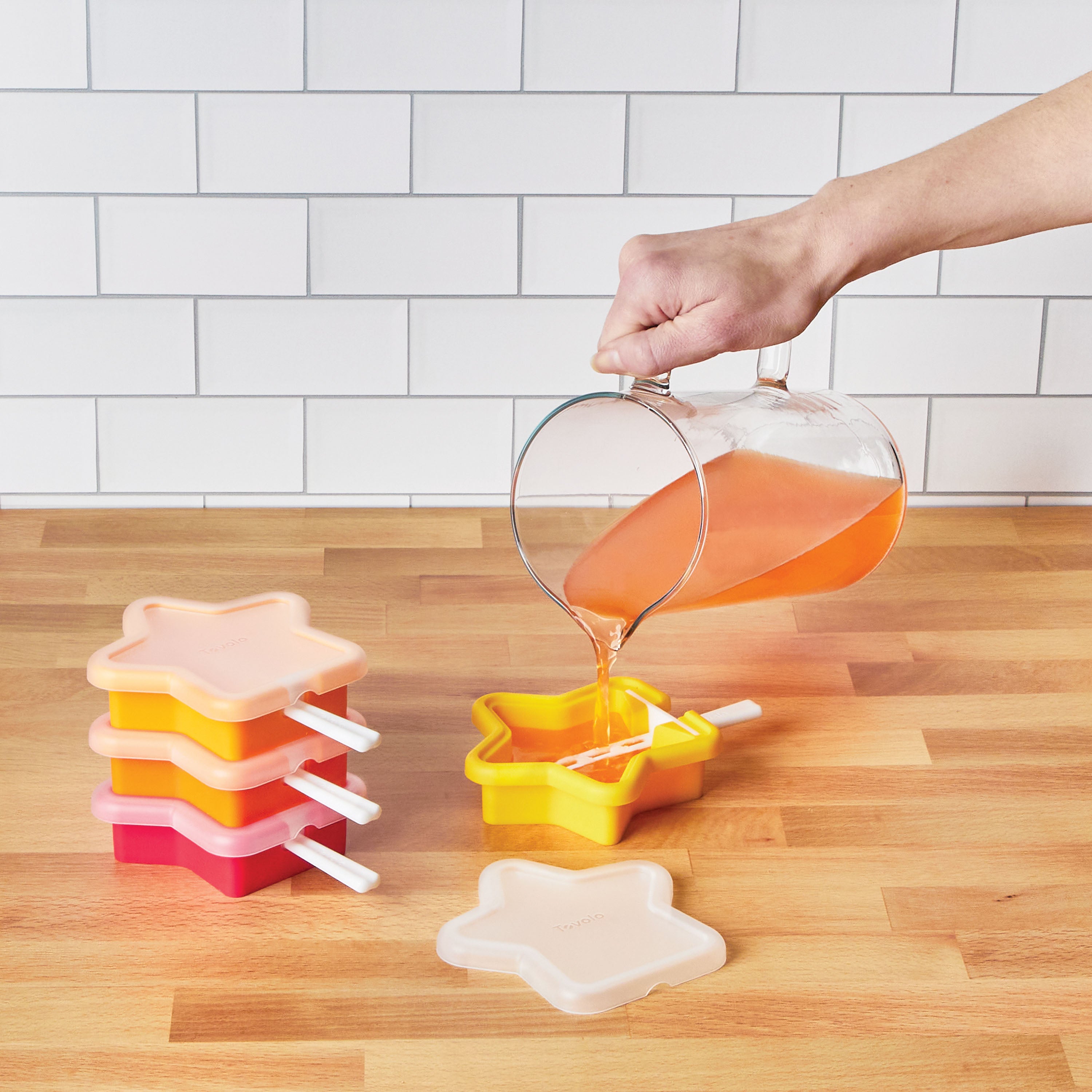 A hand pours orange liquid from a glass pitcher into a star-shaped Pop Molds Stackable silicone popsicle maker on a wooden countertop, with other colorful molds nearby and a white tile backsplash in the background.