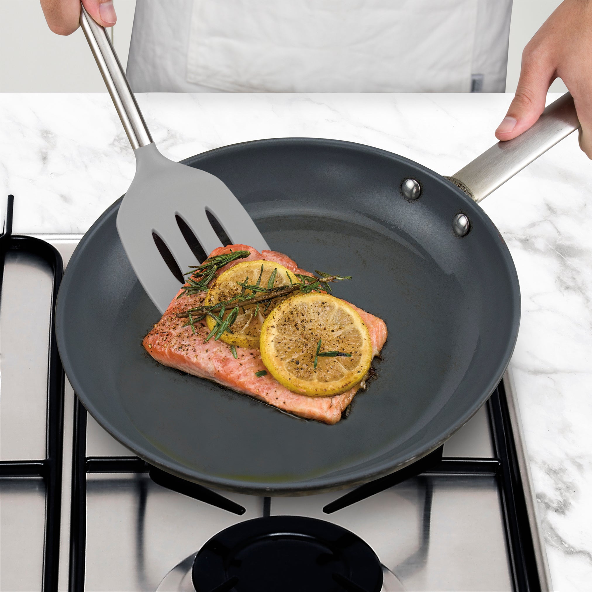 A person cooks a salmon fillet with lemon slices and herbs in a non-stick frying pan on a gas stove, using the Silicone Slotted Turner.