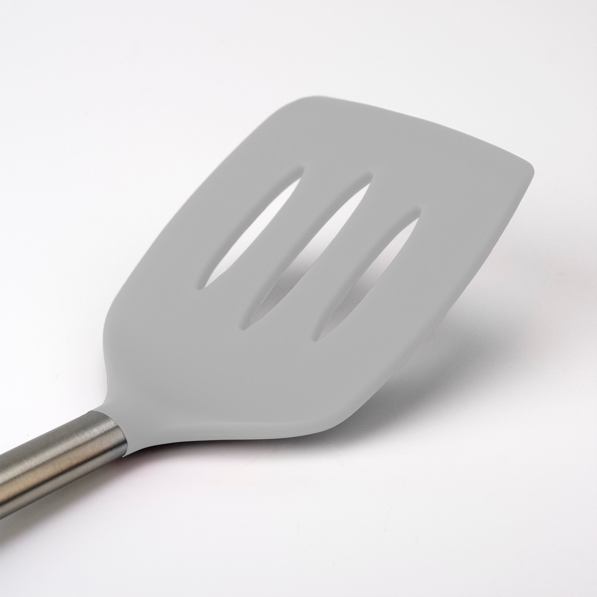 A close-up of the Silicone Slotted Turner shows its gray silicone head and stainless steel handle, safe for nonstick cookware, placed on a white background.