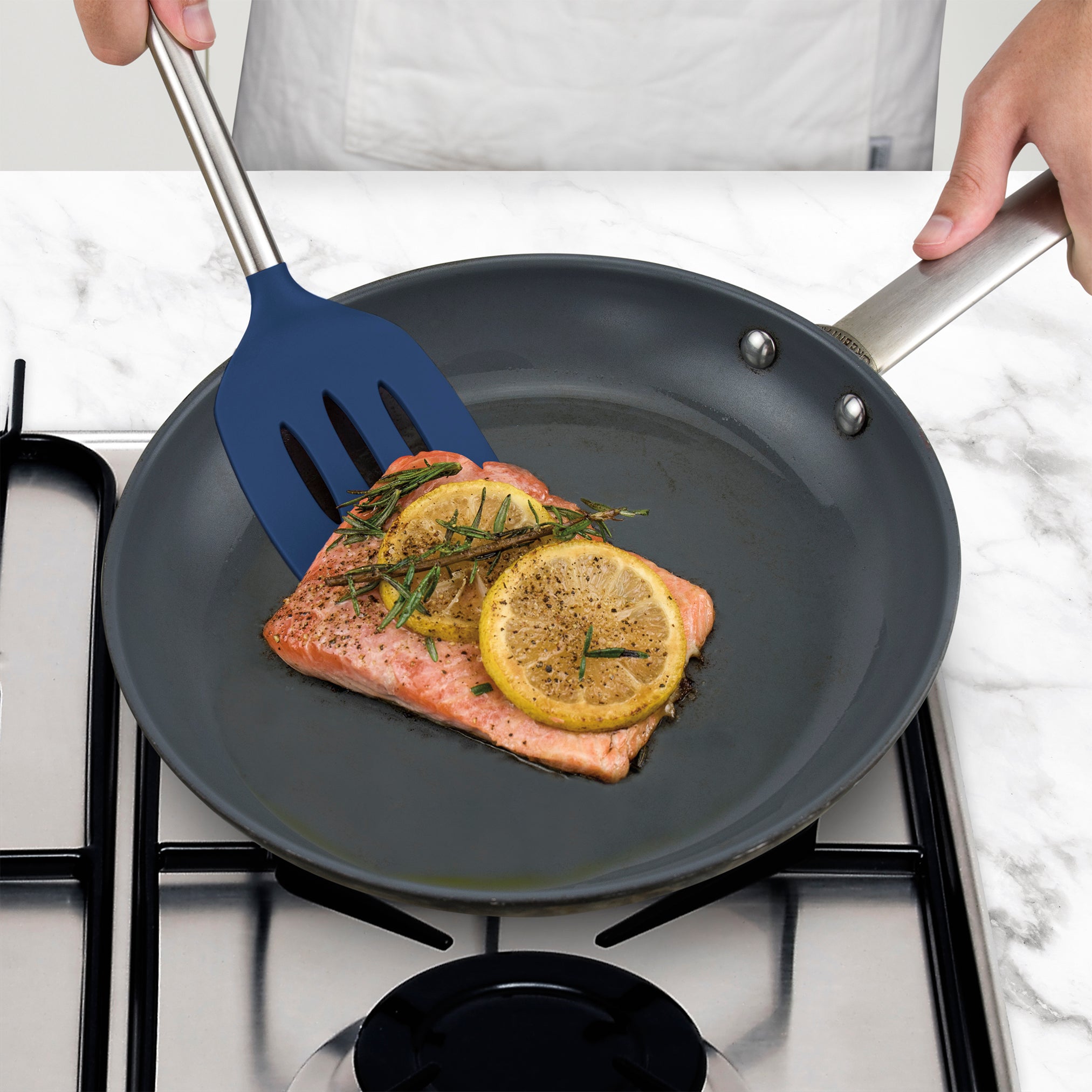 A person uses a Silicone Slotted Turner to cook a salmon fillet topped with lemon slices and herbs in a frying pan on a gas stove.