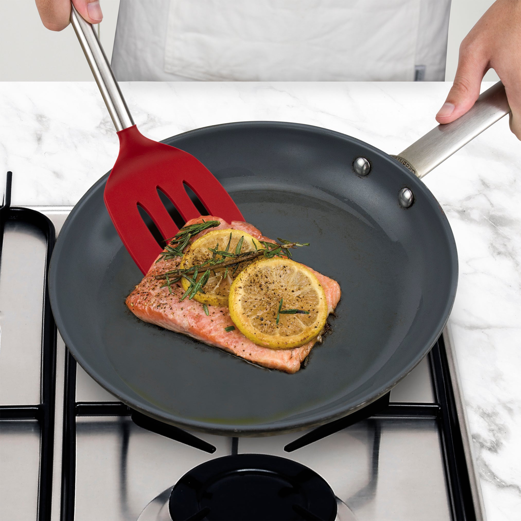 A person cooks a piece of salmon with lemon and herbs in a frying pan on a gas stove using a Silicone Slotted Turner.
