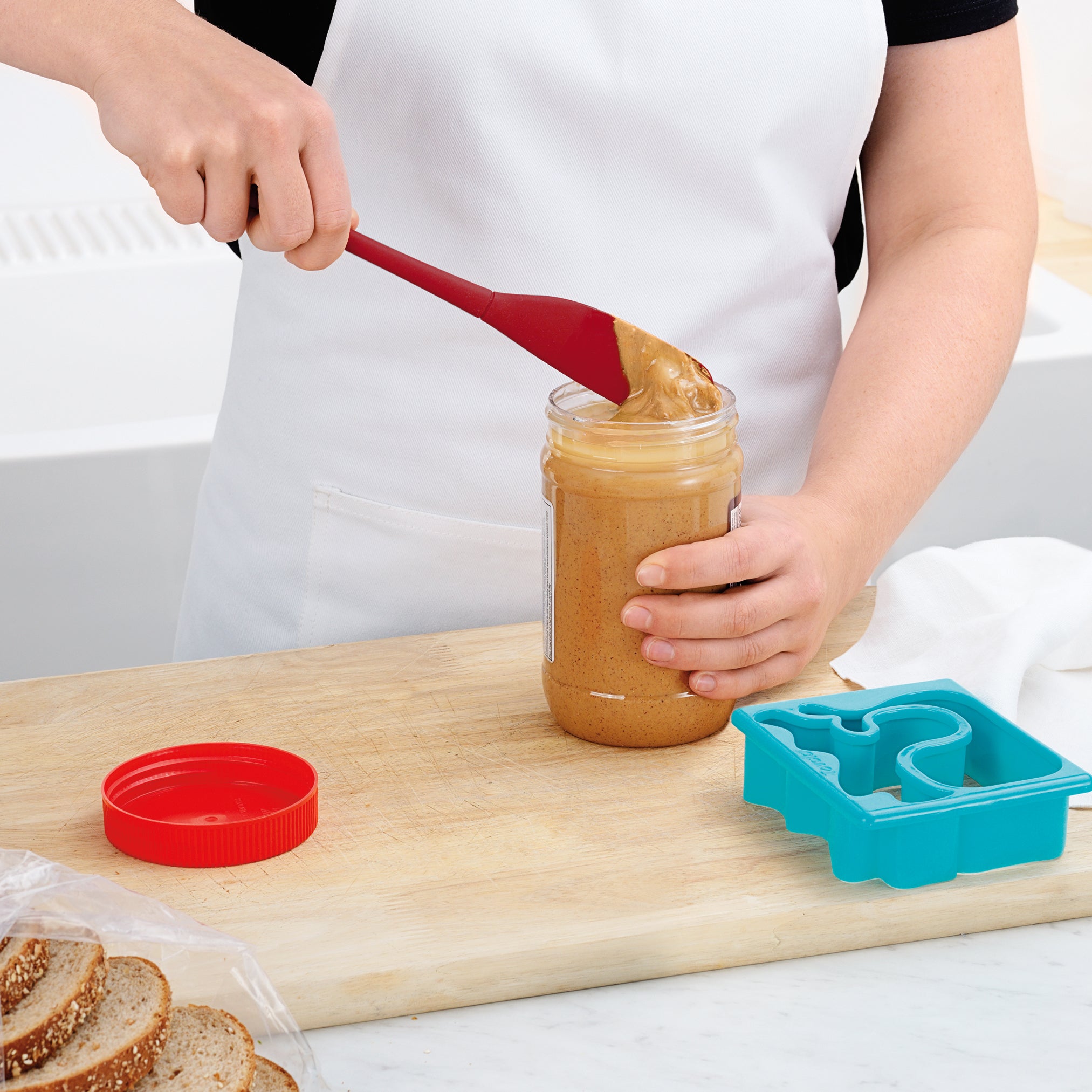 Wearing a white apron, someone uses the Flex-Core Jar Scraper—a red silicone tool—to scoop peanut butter from a jar on a wooden cutting board, with slices of bread and a blue sandwich cutter nearby. It ensures every bit is scraped out.