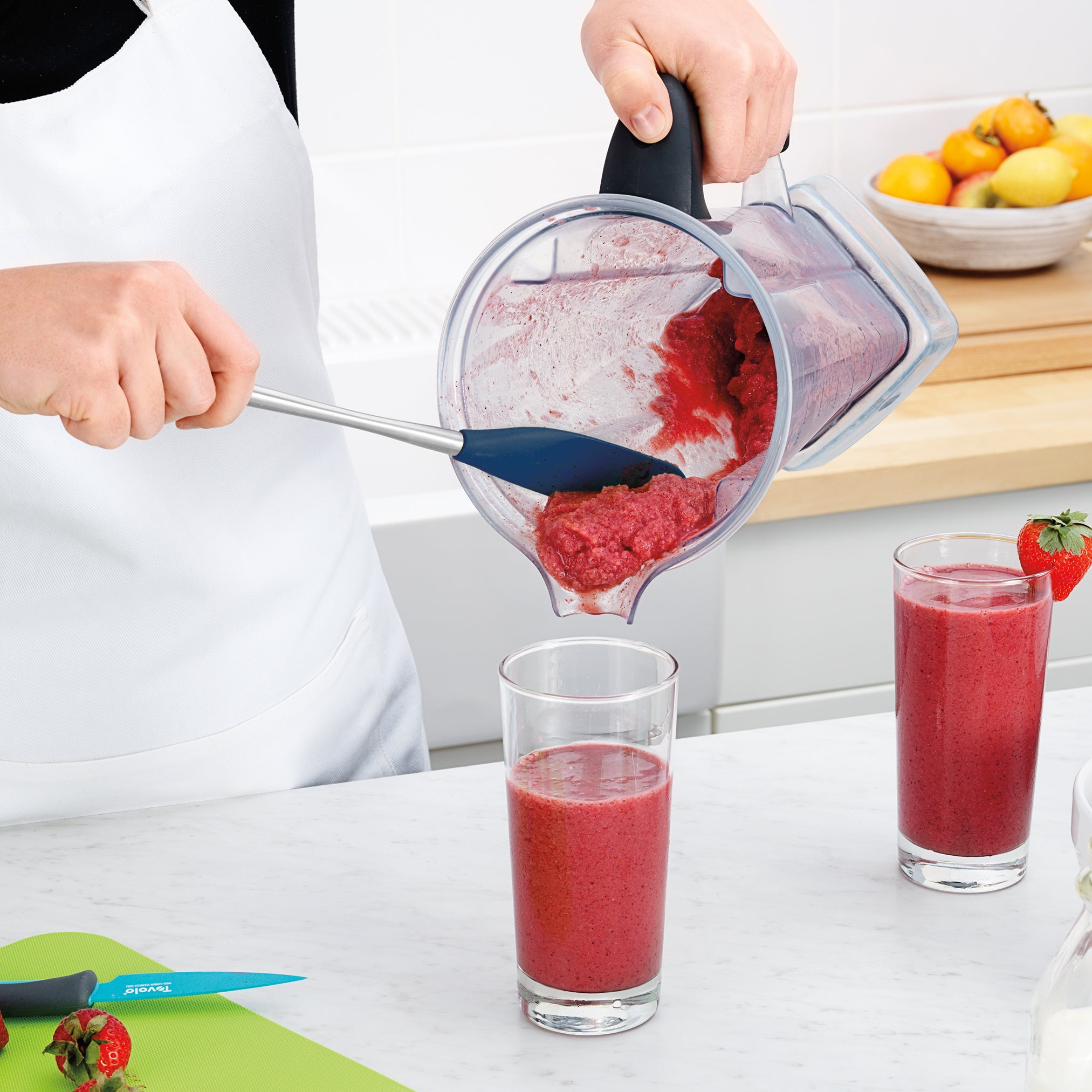 Wearing a white apron, a person uses the Flex-Core Jar Scraper to pour a thick red smoothie from a blender into a glass on the kitchen counter, surrounded by another glass, strawberries, a knife, cutting board, and fruit bowl.