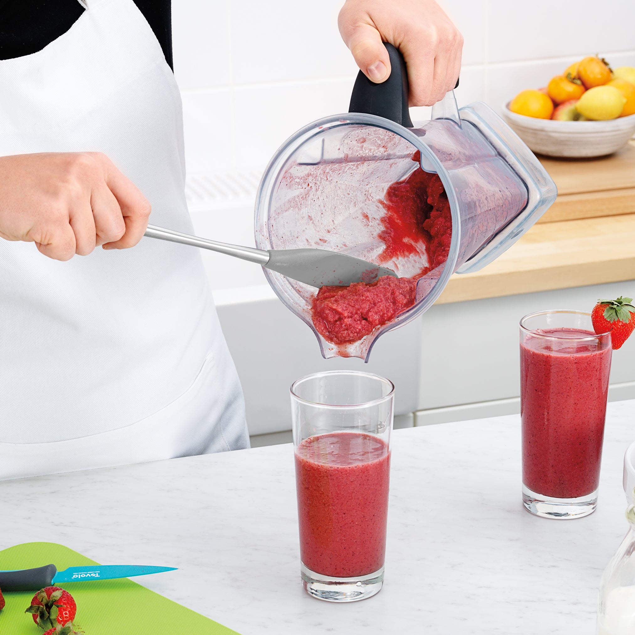 Wearing a white apron, a person uses the Flex-Core Jar Scraper to pour a thick red smoothie from a blender into a glass. Another smoothie glass, plus a green cutting board with kitchen tools, sits on the counter.