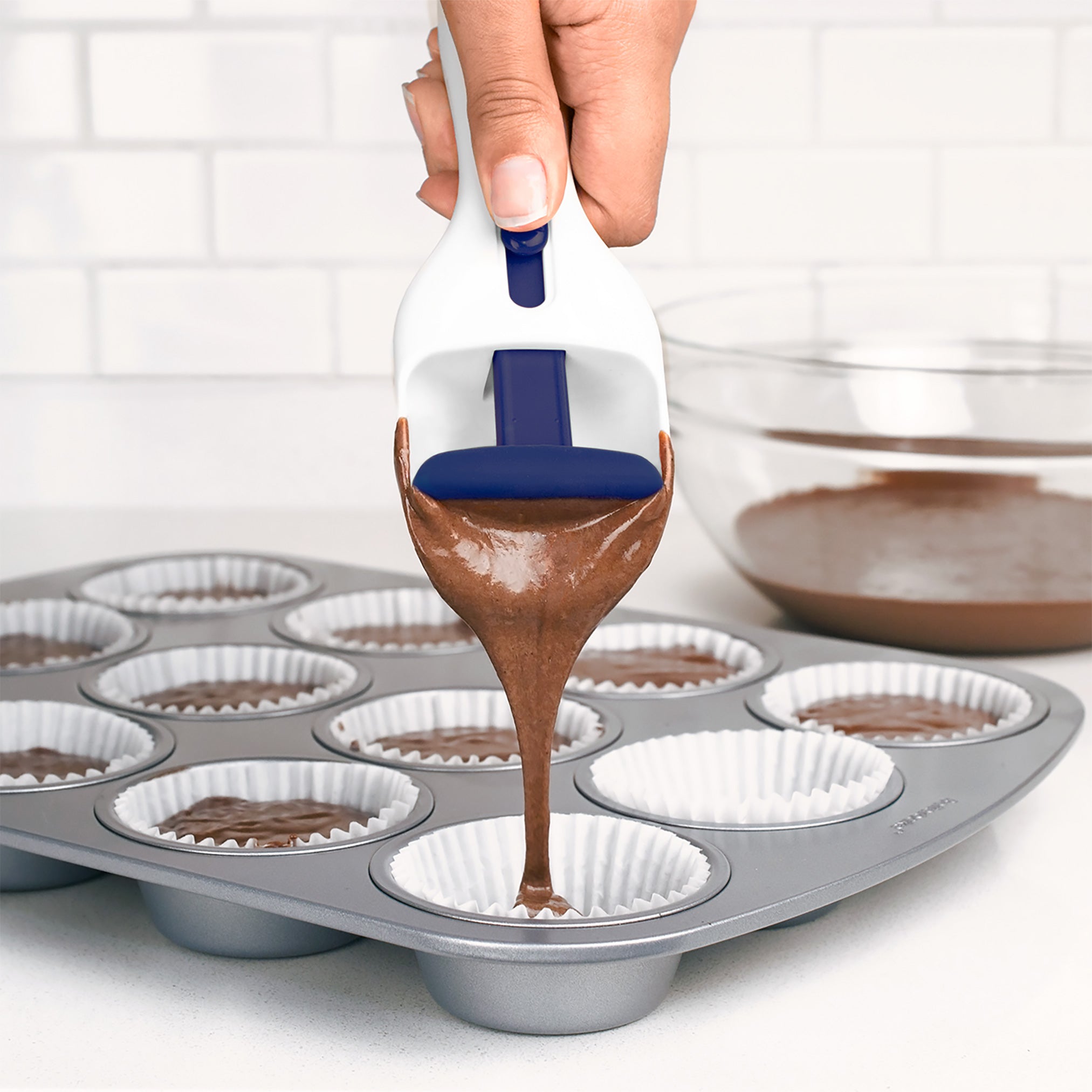 A hand uses the Cupcake Scoop with a blue trigger to pour chocolate batter into paper-lined muffin cups on a metal tray. A glass bowl with extra batter is visible in the background.