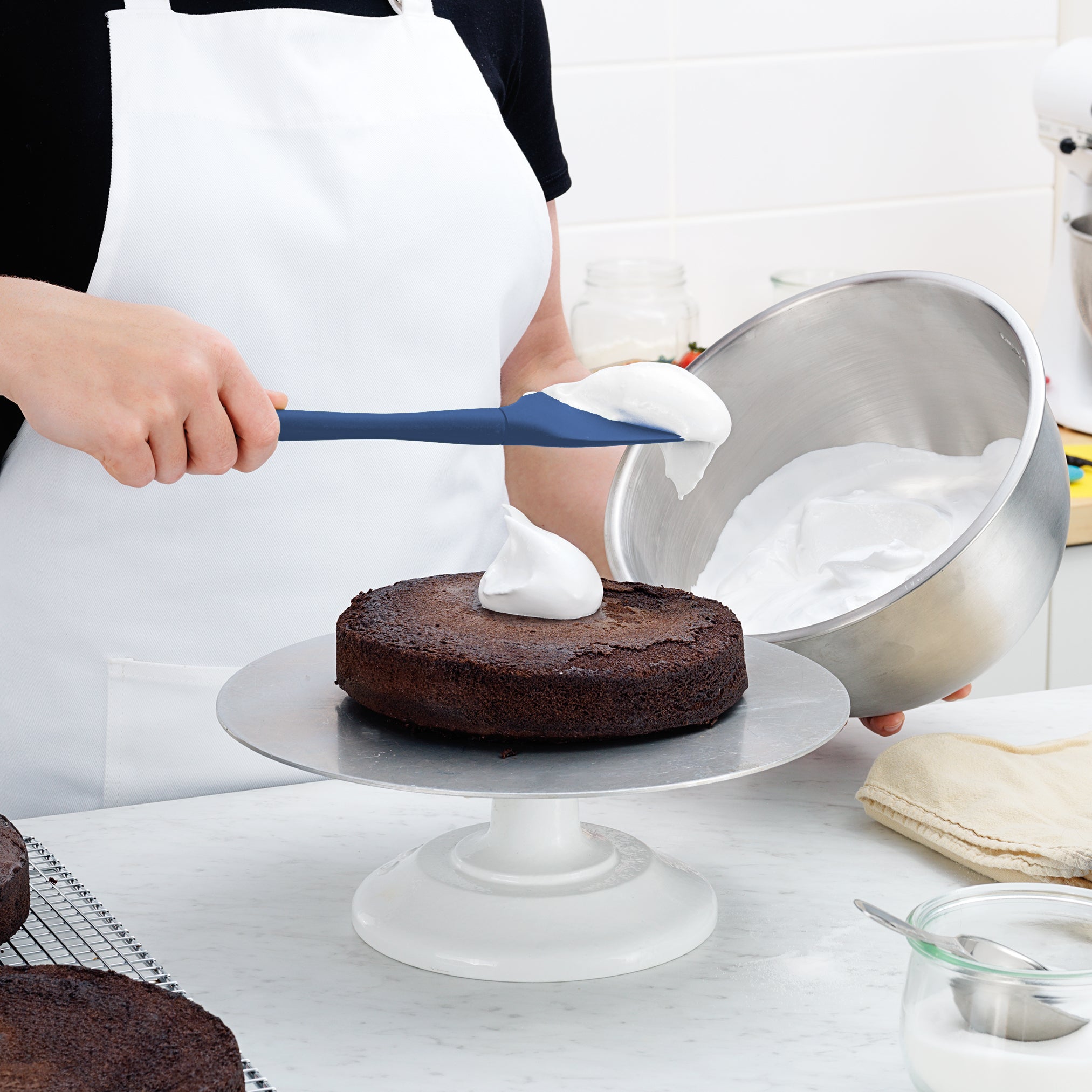 Wearing a white apron, a person spreads whipped cream on a chocolate cake using the Flex-Core Spoonula. The cake rests on a rotating stand, while the other hand holds a metal bowl of cream.