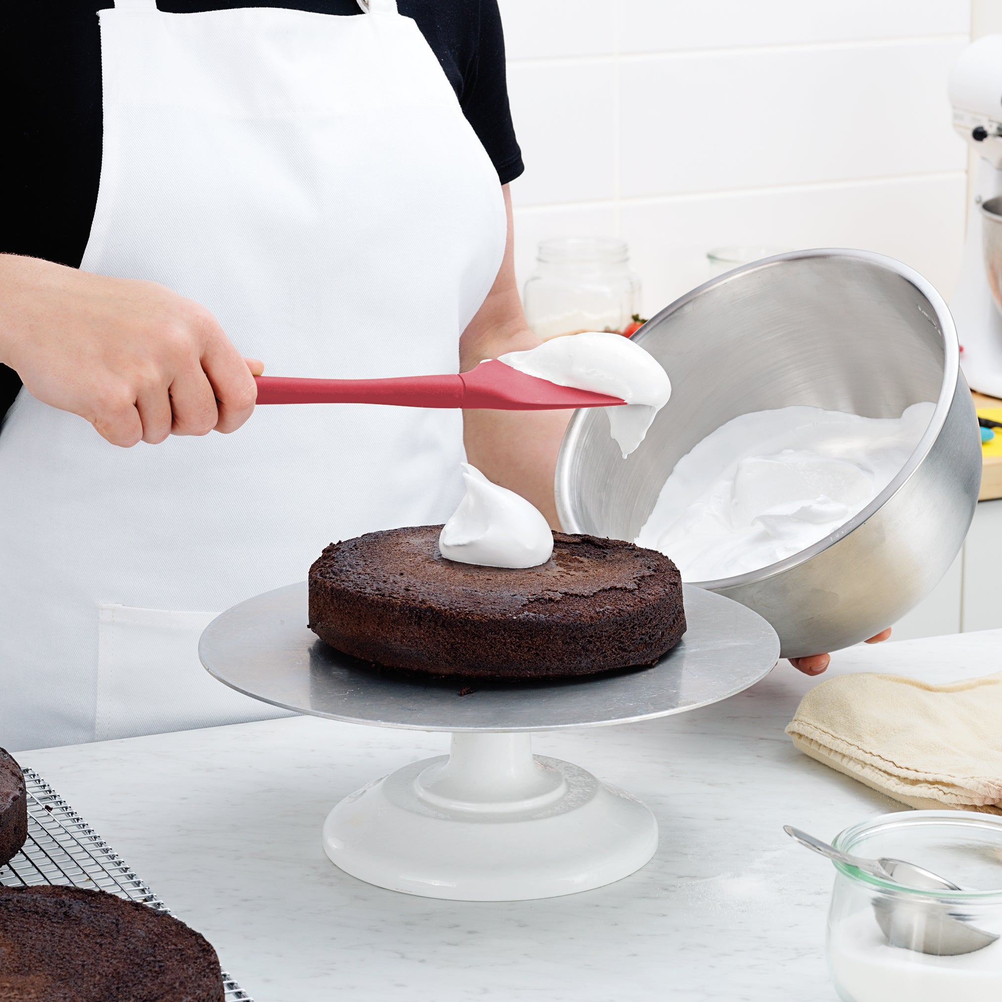 Wearing a white apron, a person uses the Flex-Core Spoonula to spread white frosting on a round chocolate cake, which rests on a stand. They hold a large metal mixing bowl in the other hand.