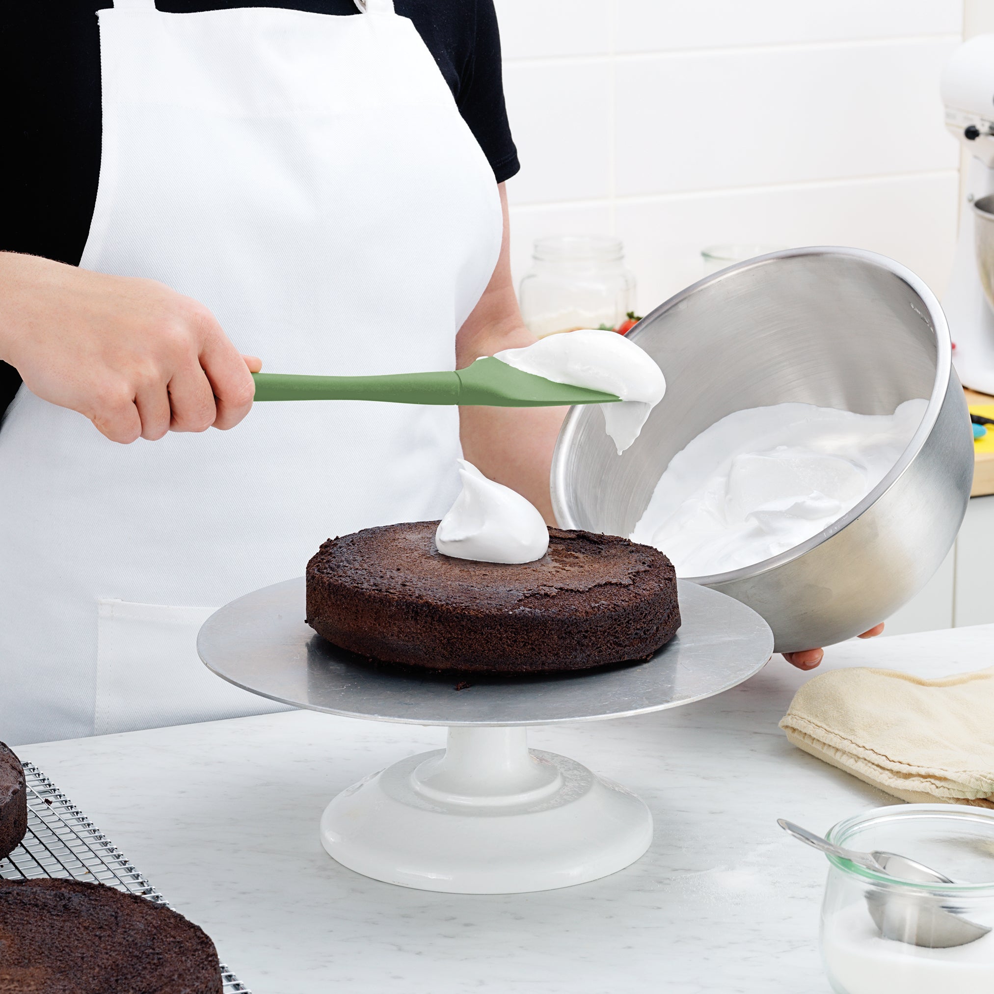 Wearing a white apron in a bright, clean kitchen, a person uses the Flex-Core Spoonula to spread white frosting onto a chocolate cake on a rotating stand while holding a metal bowl of frosting.