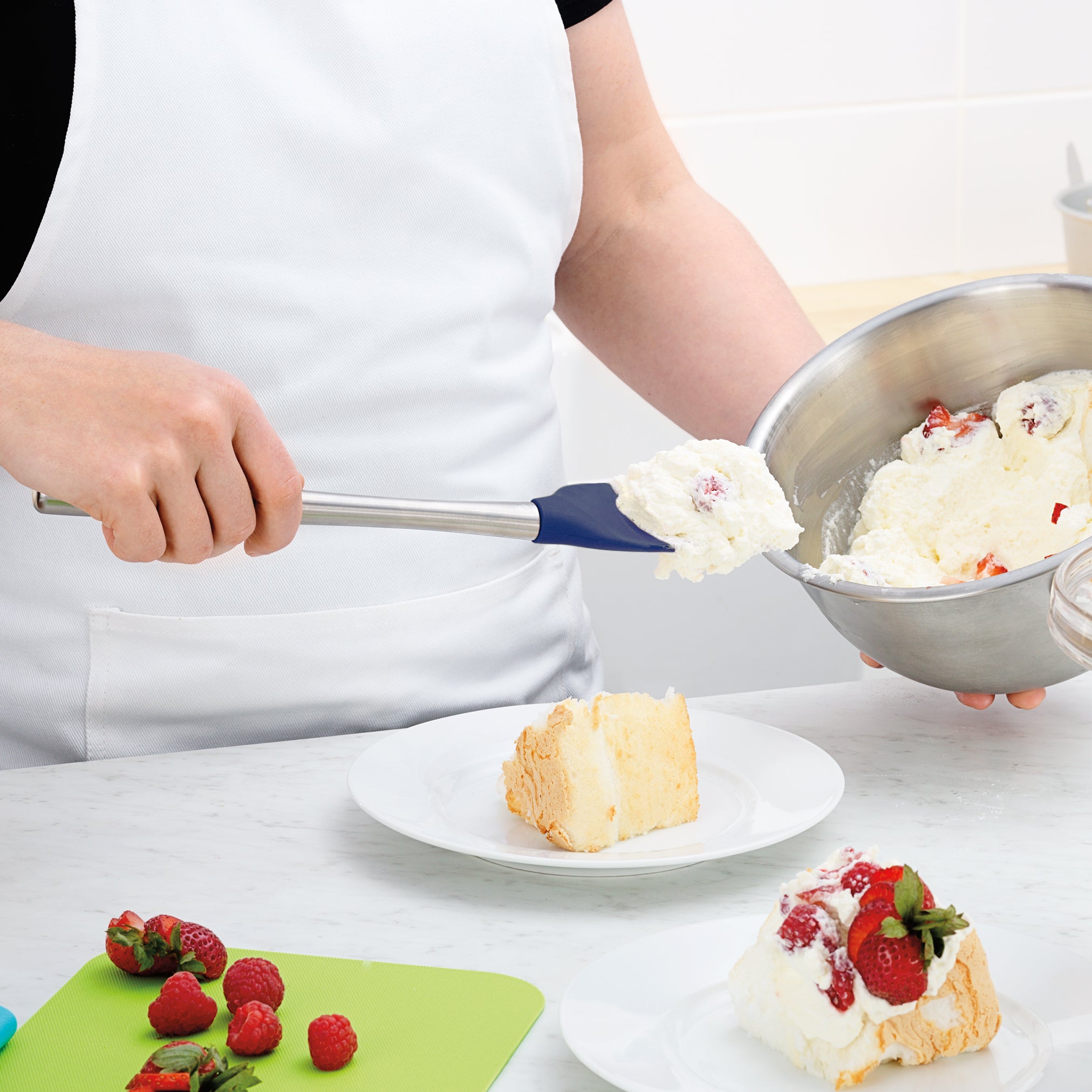 Wearing a white apron, a person uses the Flex-Core Spoonula to scoop whipped cream with strawberries from a metal bowl onto a cake slice; sliced strawberries and raspberries sit on a green cutting board nearby.