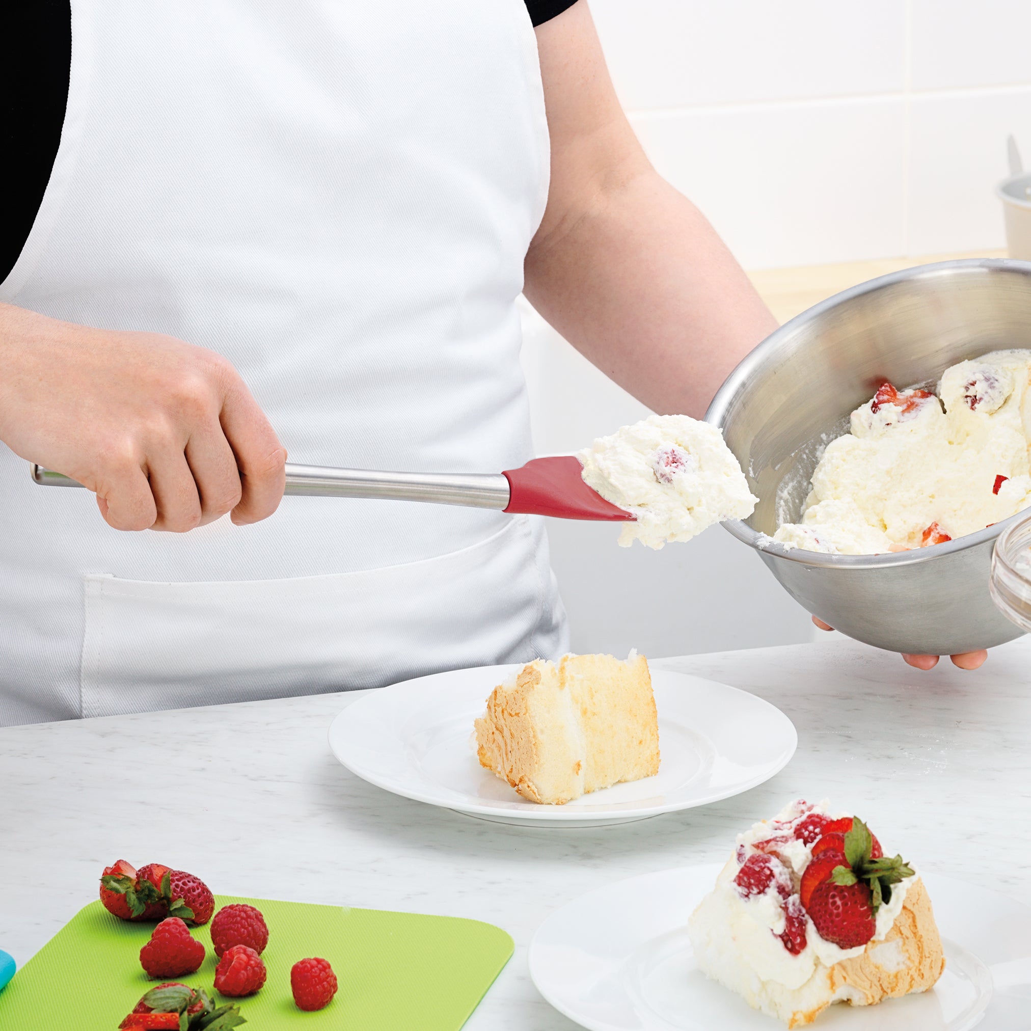 Wearing a white apron, a person uses the Flex-Core Spoonula to add whipped cream and berries to cake. Fresh strawberries and raspberries are on a green cutting board nearby.