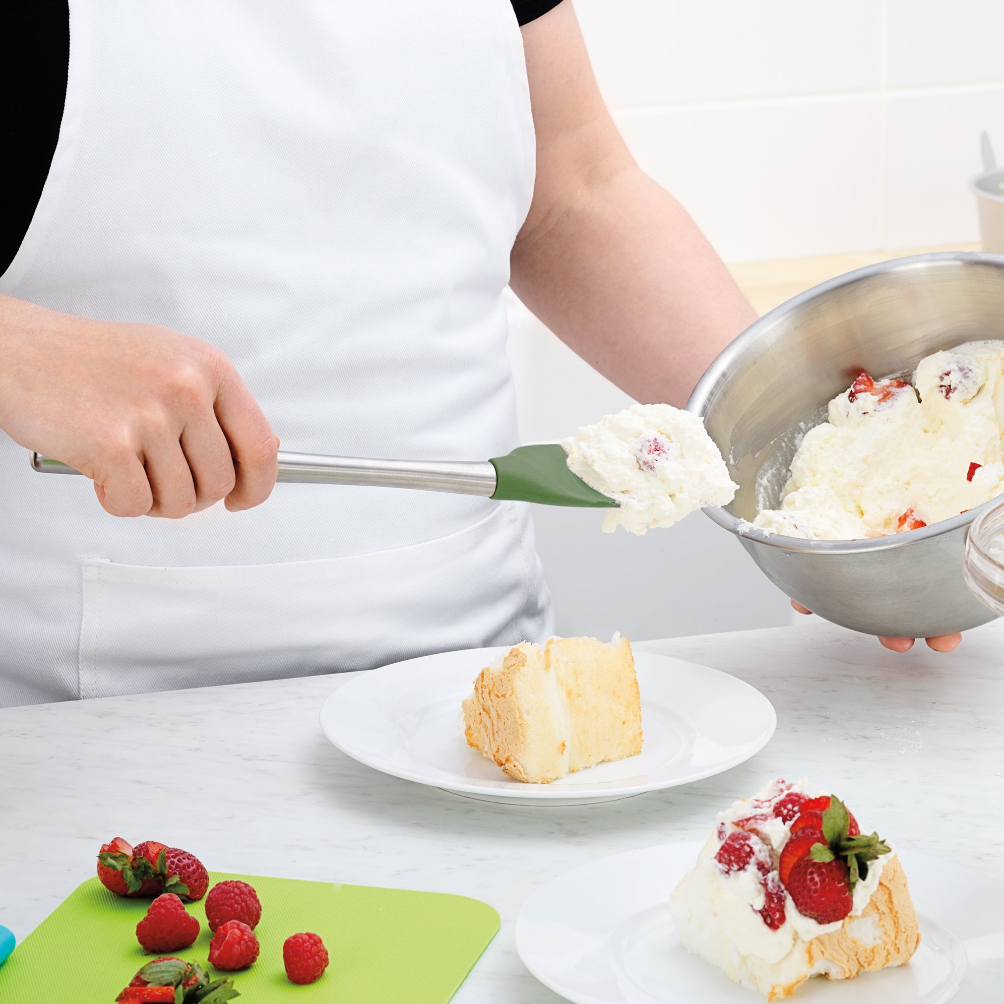 Wearing a white apron, someone uses a Flex-Core Spoonula to top a cake slice with whipped cream and strawberries, while fresh strawberries and raspberries sit on a green cutting board nearby.
