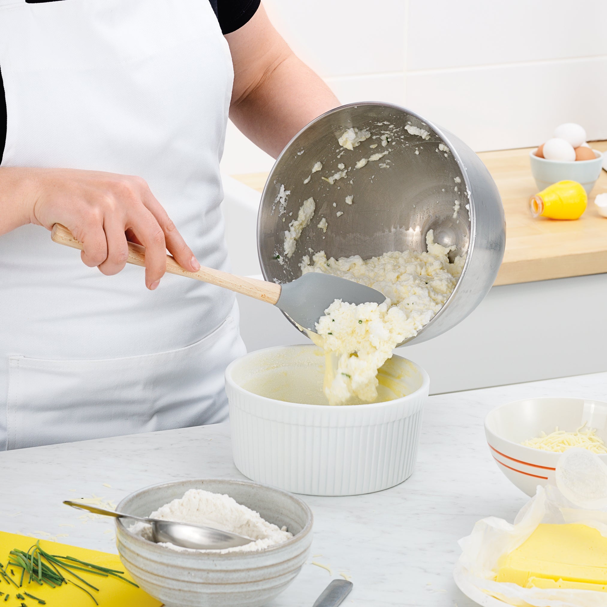 Wearing a white apron, someone uses the Flex-Core Spoonula to scoop a creamy mixture from a metal bowl into a round white baking dish, with flour, butter, and cheese visible on the kitchen counter.
