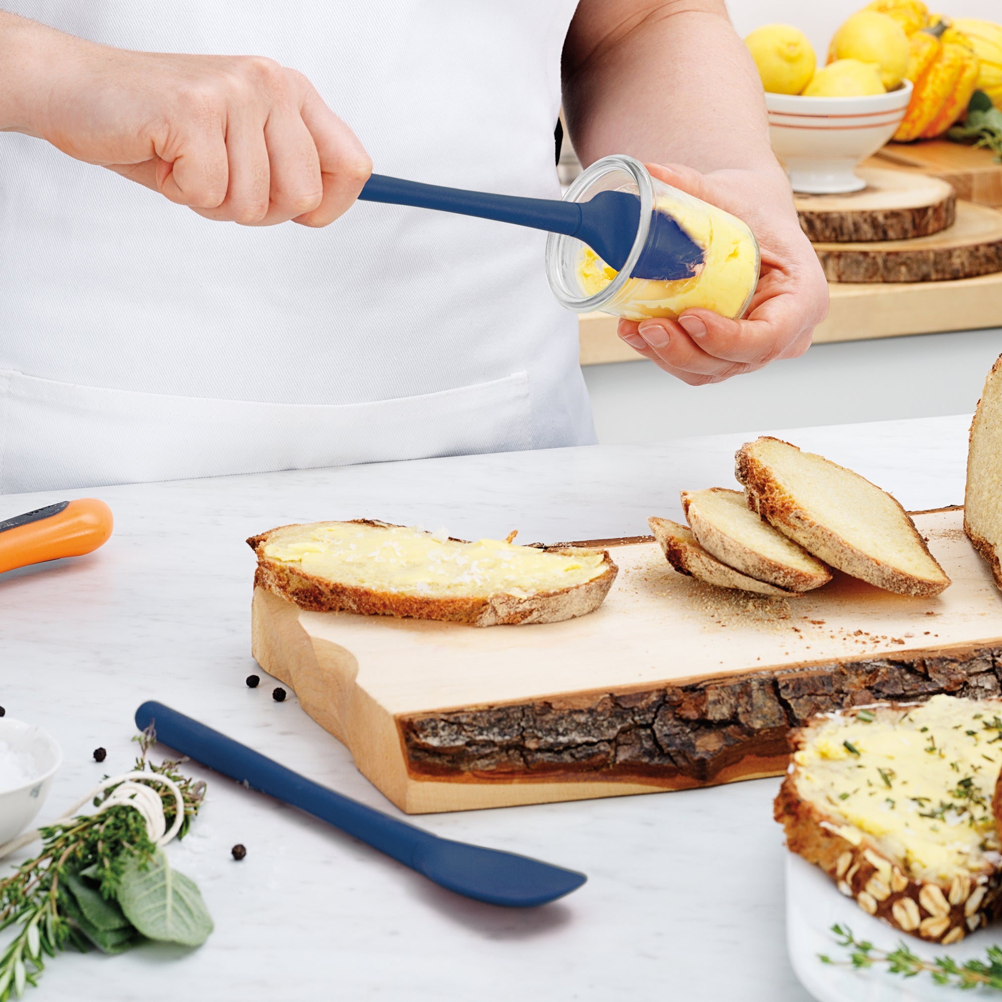 Using the Flex-Core Mini Spat + Spoonula, someone spreads yellow butter on rustic bread slices. Fresh herbs, black pepper, and lemons sit on a white kitchen counter. The spatula is blue and made of silicone.