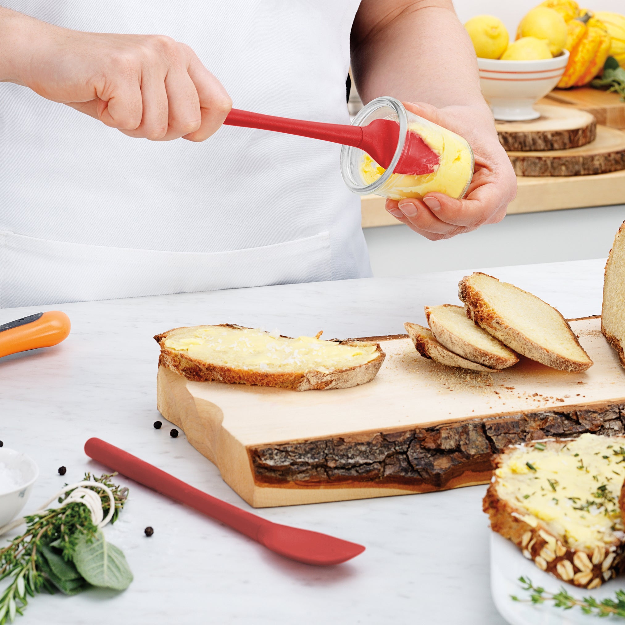 In a bright kitchen, someone spreads yellow butter on rustic bread slices with the Flex-Core Mini Spat + Spoonula. Fresh herbs, peppercorns, and lemons on the counter complete the fresh, inviting atmosphere.