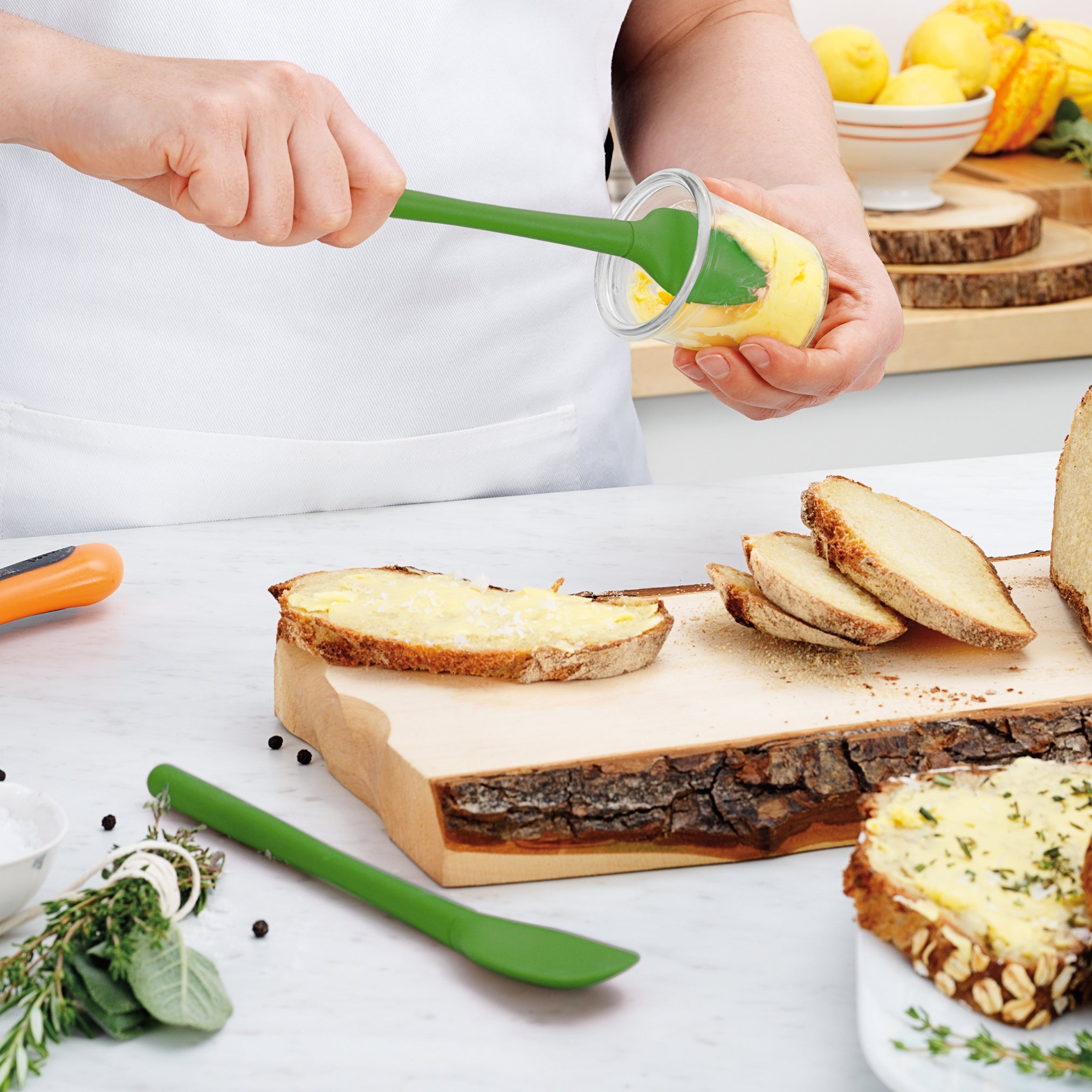Using the Flex-Core Mini Spat + Spoonula, a person spreads yellow butter from a small glass jar onto rustic bread slices on a wooden cutting board, with fresh herbs and a green kitchen utensil on the white countertop.