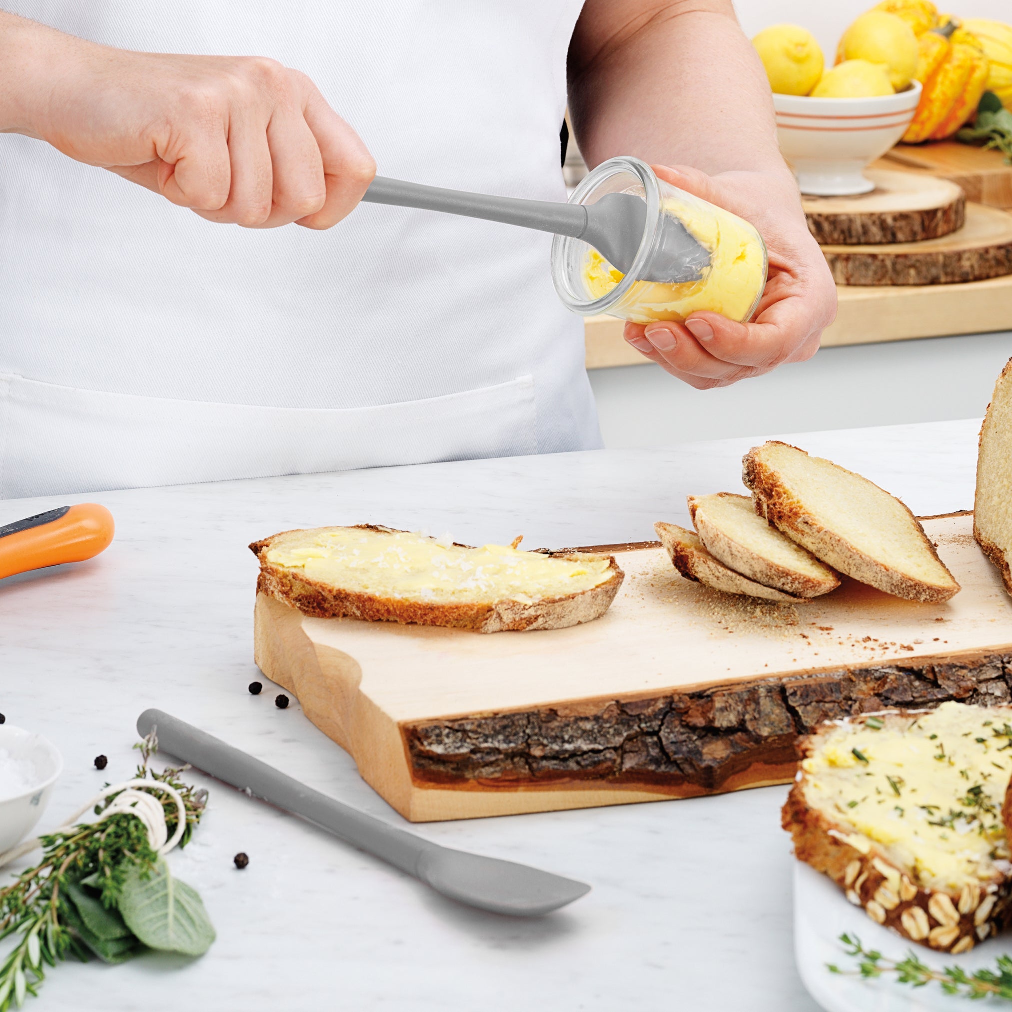 Using the Flex-Core Mini Spat + Spoonula, someone spreads yellow butter from a glass jar onto rustic bread slices on a wooden cutting board. Fresh herbs and a heat-resistant kitchen tool sit on the white countertop nearby.