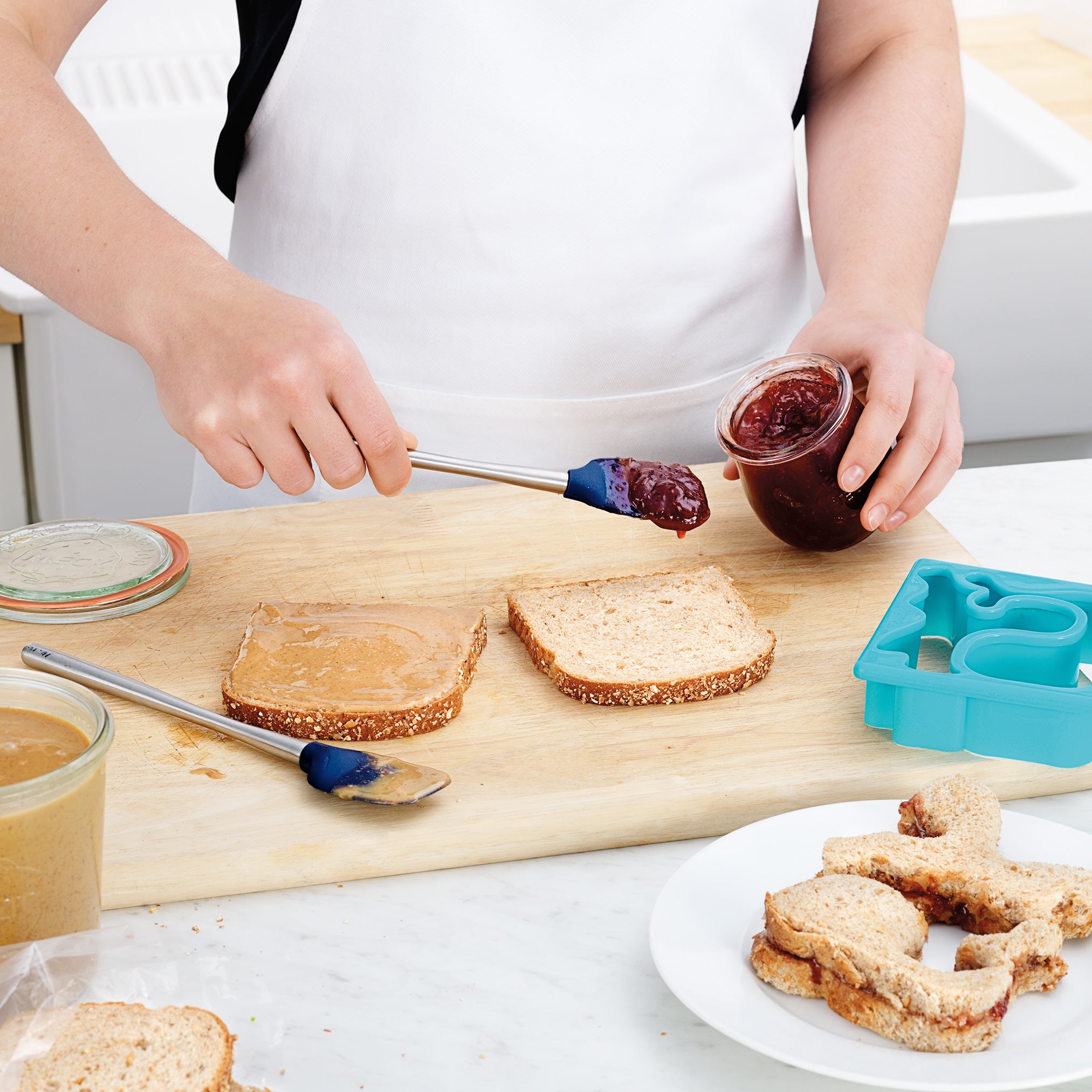A person uses the Flex-Core Mini Spat + Spoonula to spread jelly on bread, while another slice has peanut butter. Nearby are a sandwich cutter, animal-shaped sandwiches, extra bread, and jars of peanut butter and jelly.