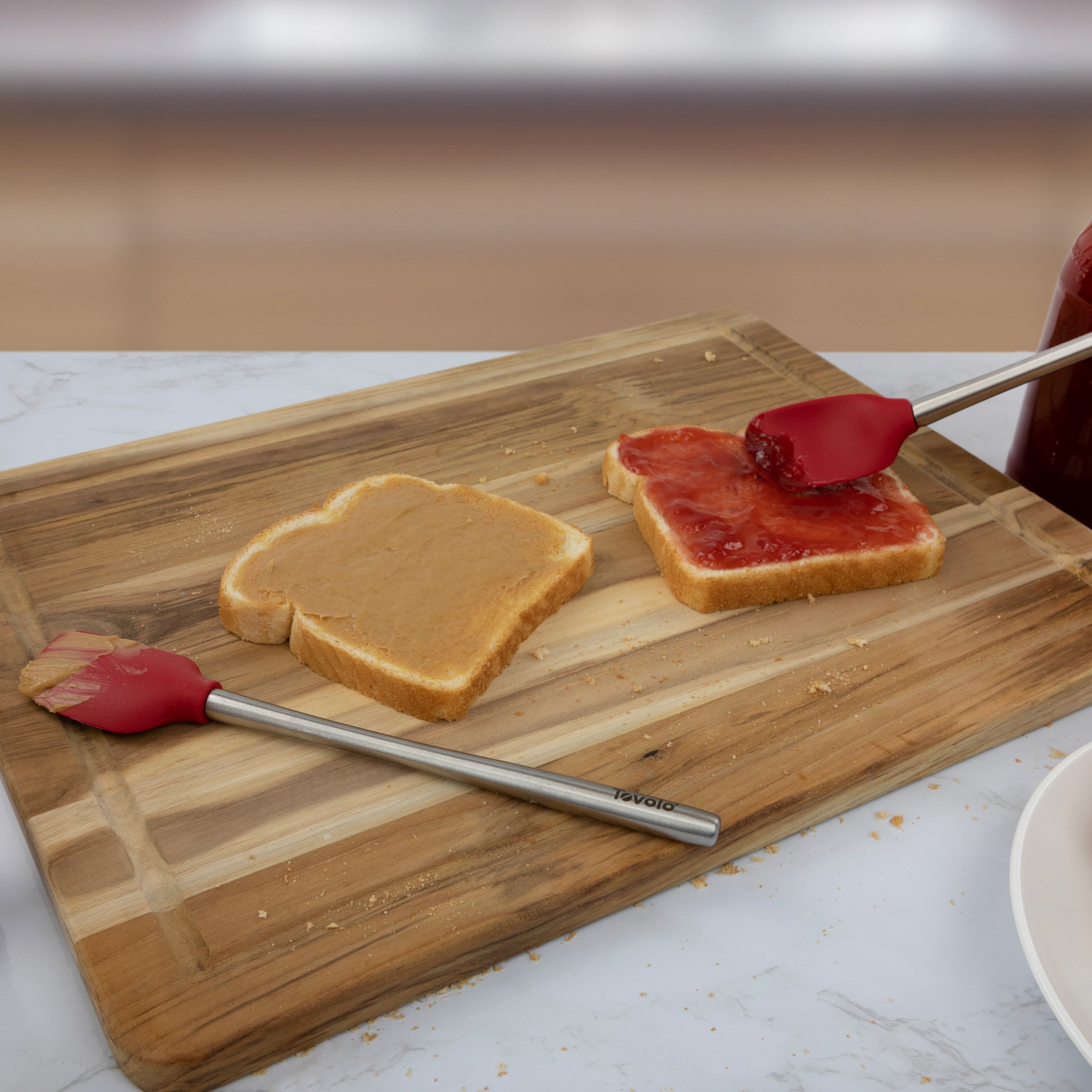 Two slices of bread on a wooden cutting board; one is topped with peanut butter, while jelly is spread on the other using the Flex-Core Mini Spat + Spoonula, a heat-resistant silicone kitchen utensil resting nearby.