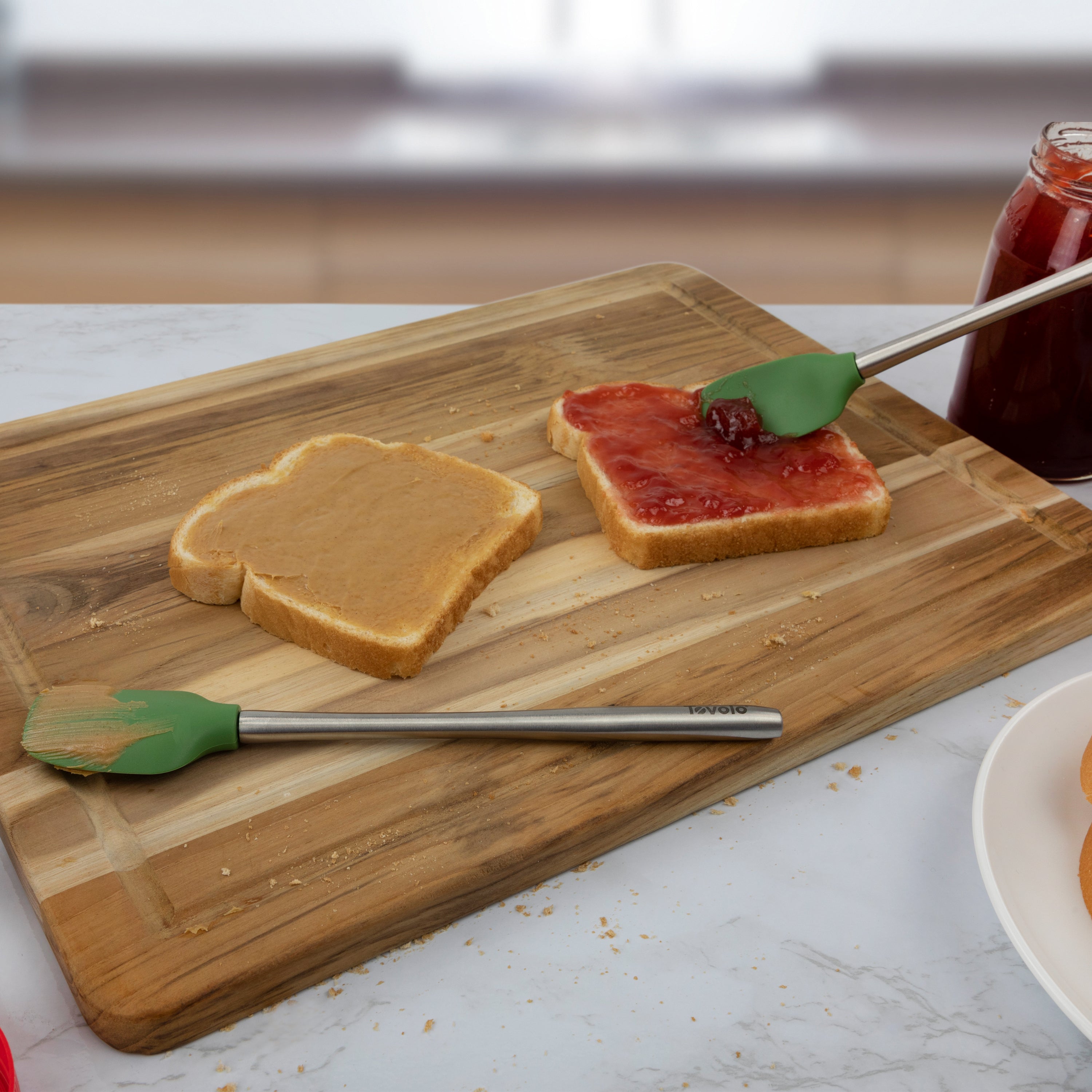 On a wooden cutting board, two bread slices—one with peanut butter, one with jelly—are shown as a green Flex-Core Mini Spat + Spoonula spreads jelly on a slice, with a jar of jelly nearby.