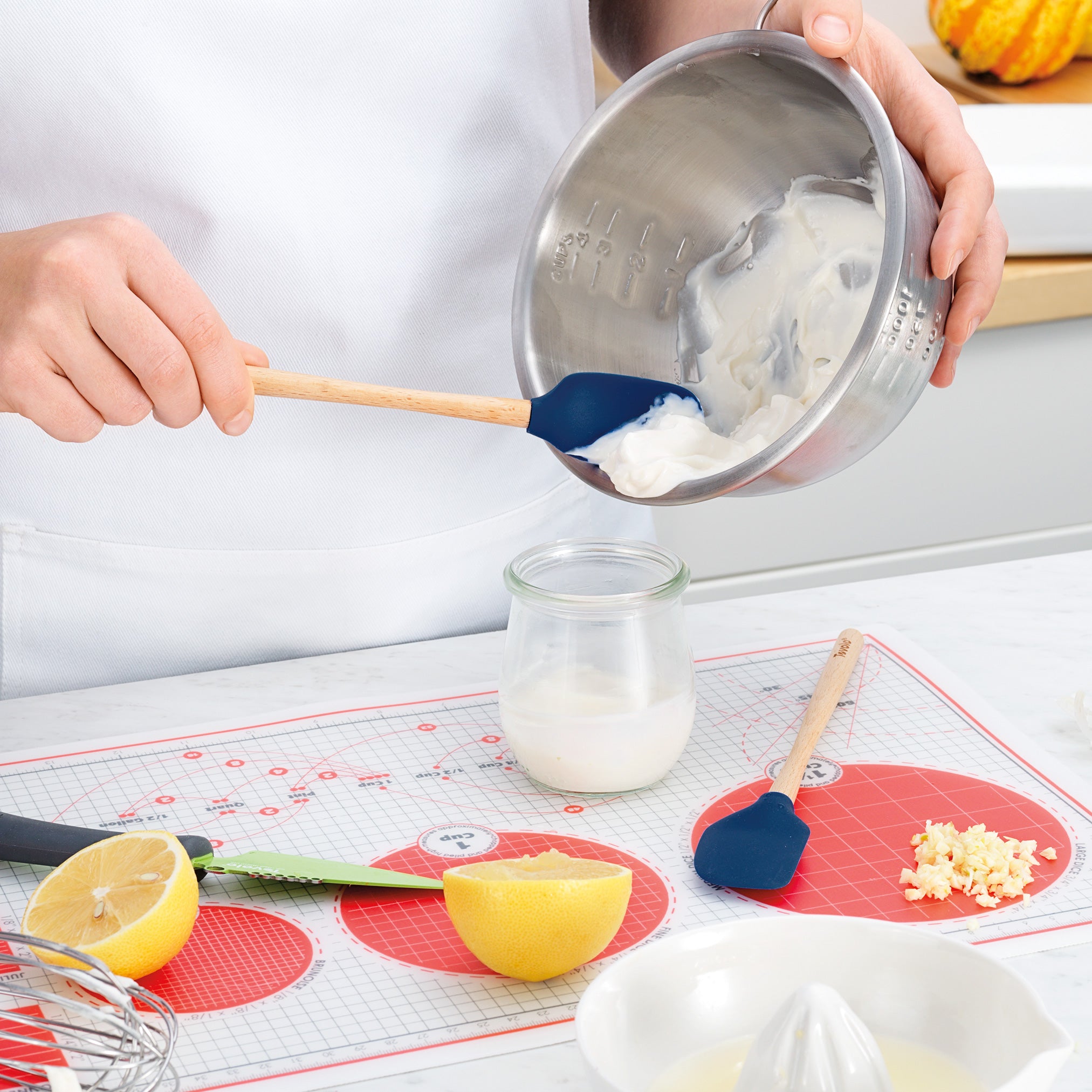 Wearing a white apron, a person uses the Flex-Core Mini Spat + Spoonula to transfer a creamy mixture from a metal bowl into a glass jar, surrounded by chopped garlic, half a lemon, and kitchen utensils on the counter.