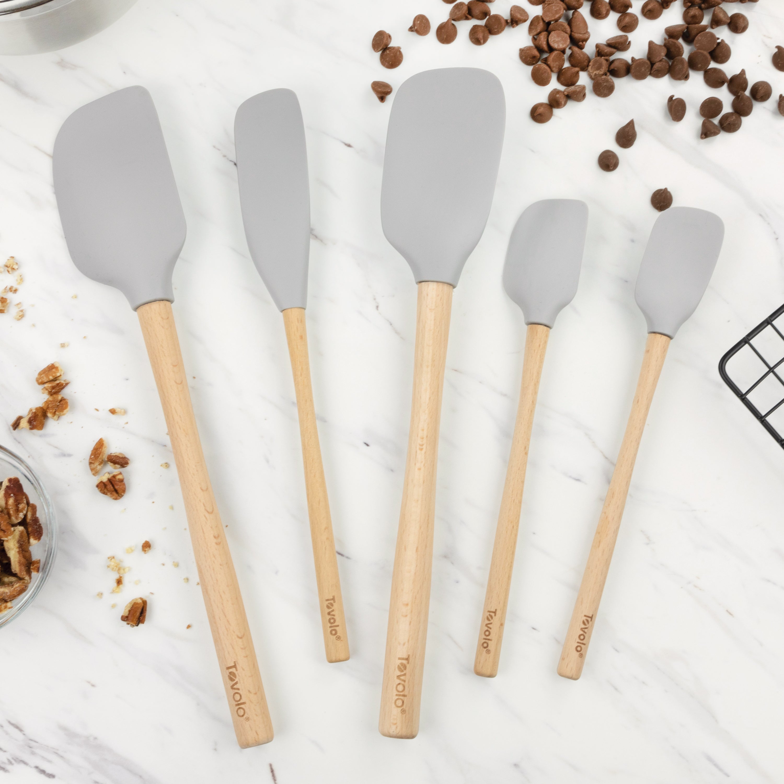 The Flex-Core Wood-Handle Spatula Set of 5 in gray, featuring assorted sizes and wooden handles, is displayed on a marble surface near chocolate chips, pecans, and a cooling rack.