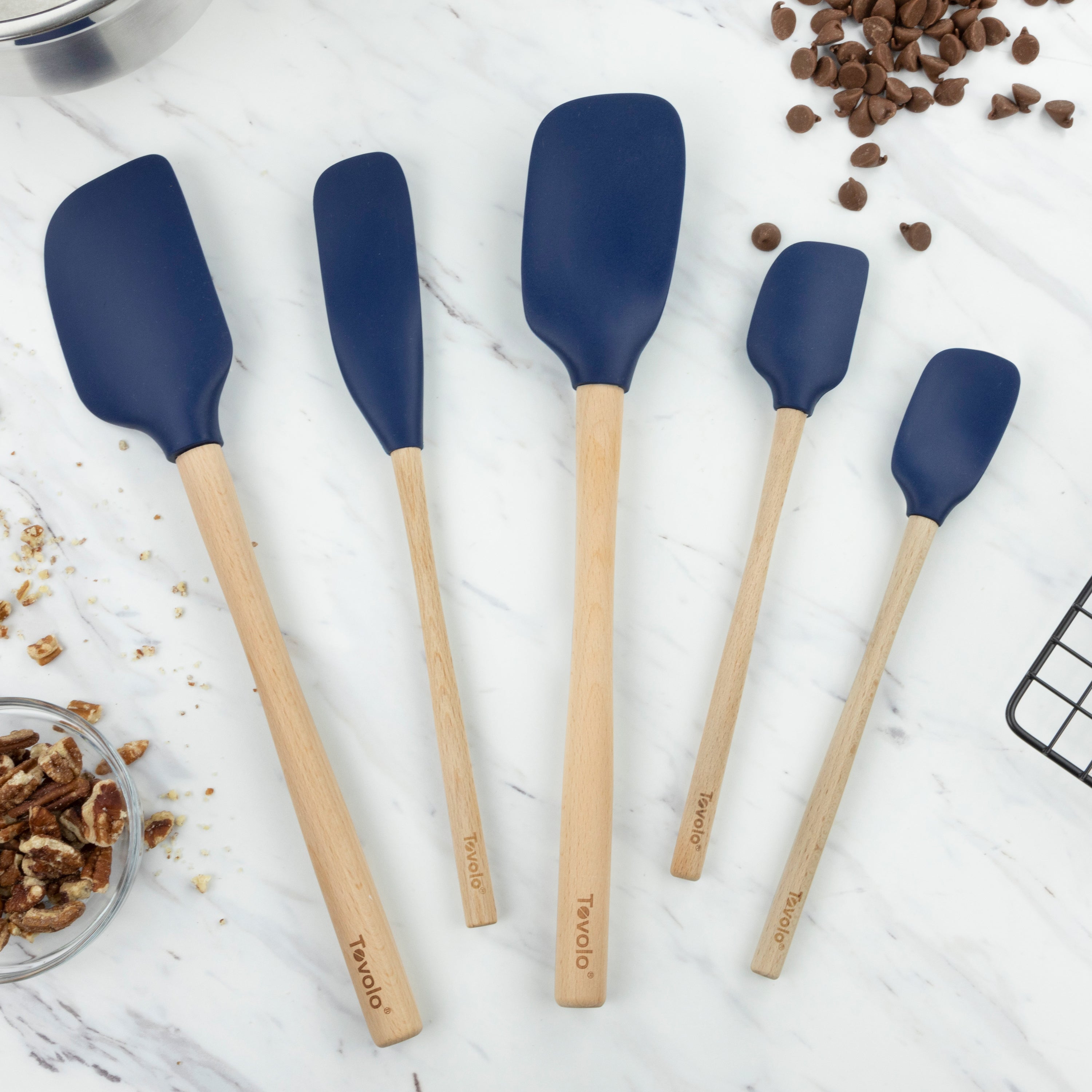 Five blue Flex-Core Wood-Handle Spatula Set of 5 pieces are arranged on a marble countertop, surrounded by chocolate chips, chopped nuts, and a cooling rack—versatile tools perfect for baking or scraping jars.