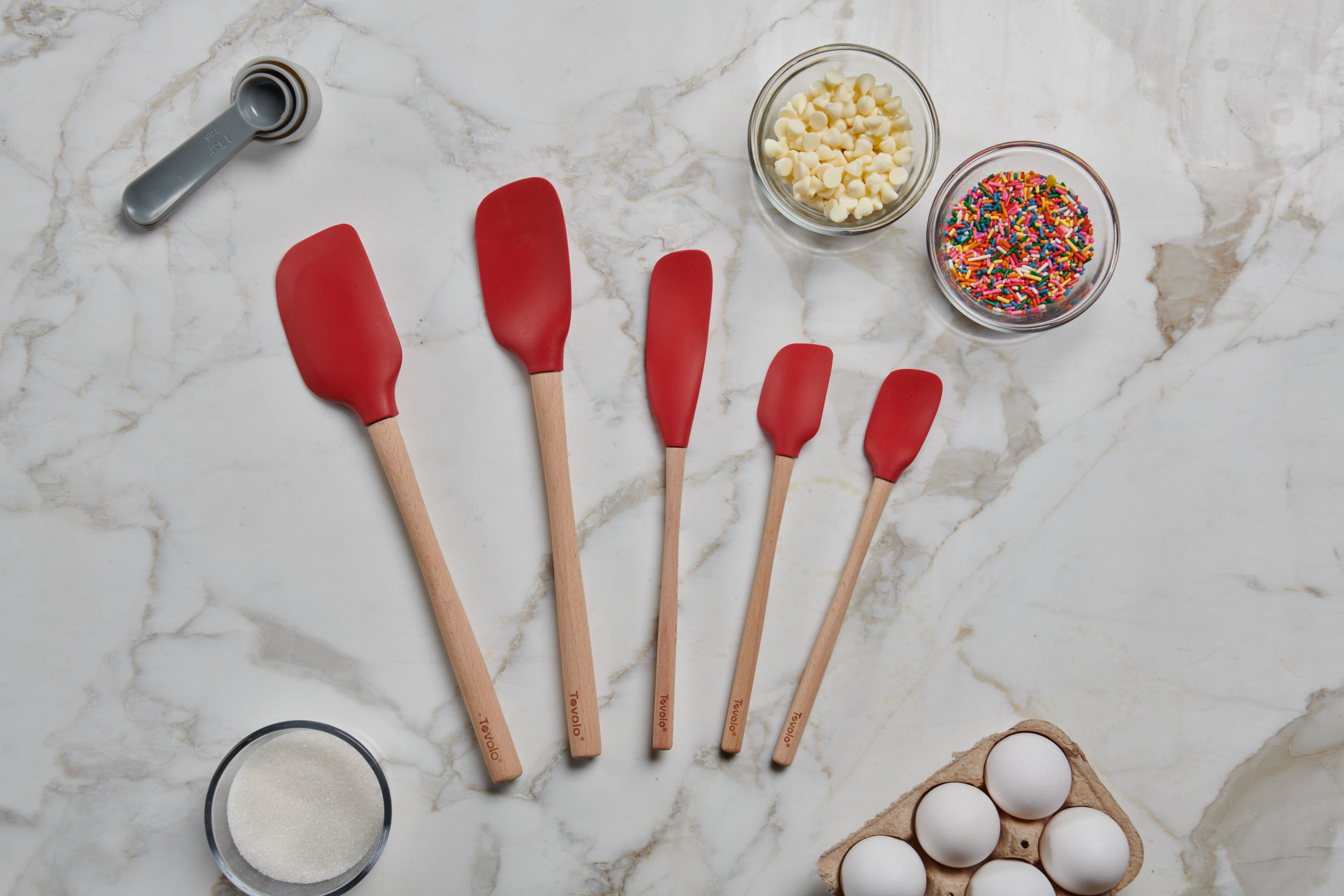 Five Flex-Core Wood-Handle Spatulas in red, each a different size, are arranged on a marble countertop with eggs, white sugar, white chocolate chips, sprinkles, and a metal tablespoon.