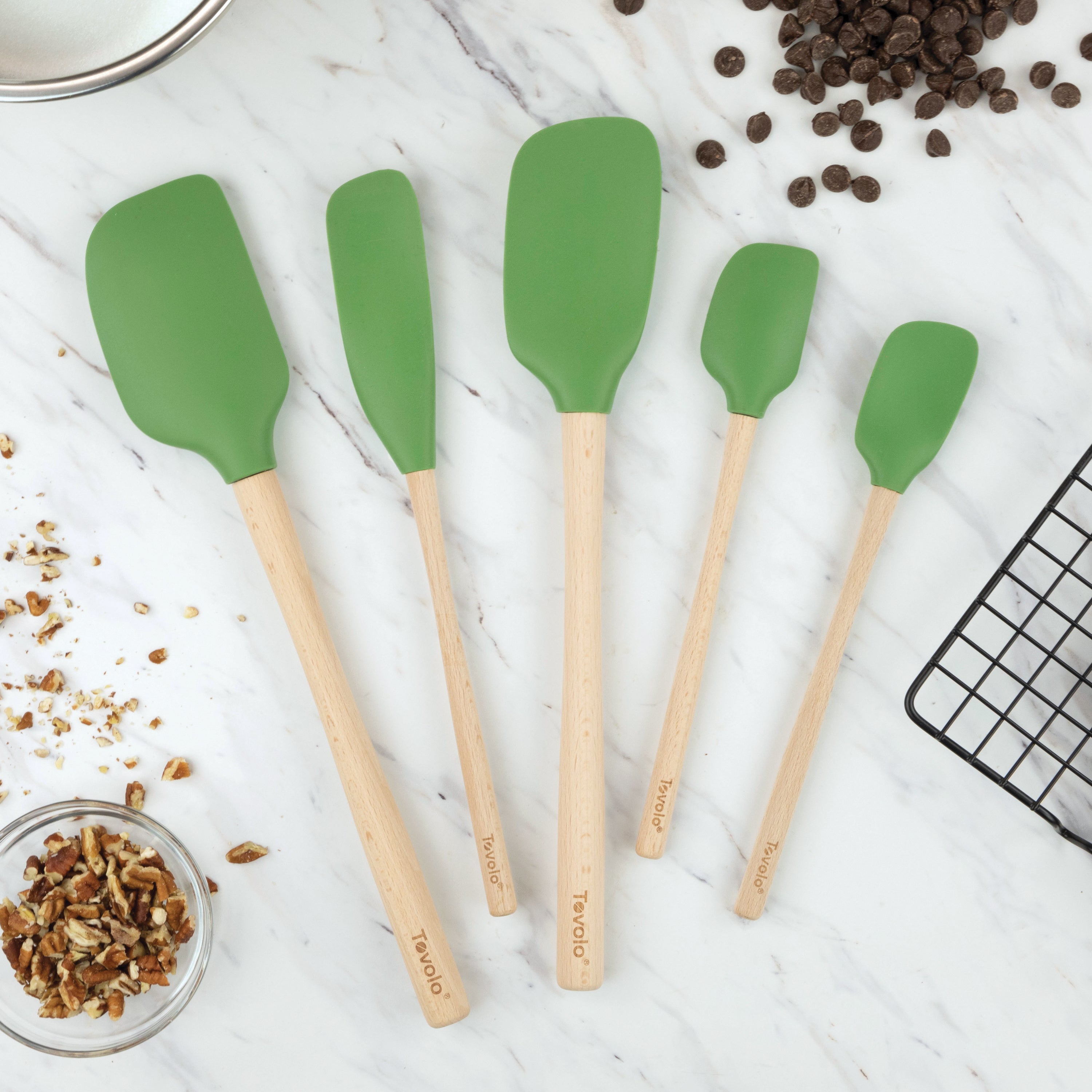 Five green Flex-Core Wood-Handle Spatula Set of 5 pieces are displayed on a white marble countertop, surrounded by chocolate chips, a bowl of chopped nuts, a metal mixing bowl, and a black cooling rack.