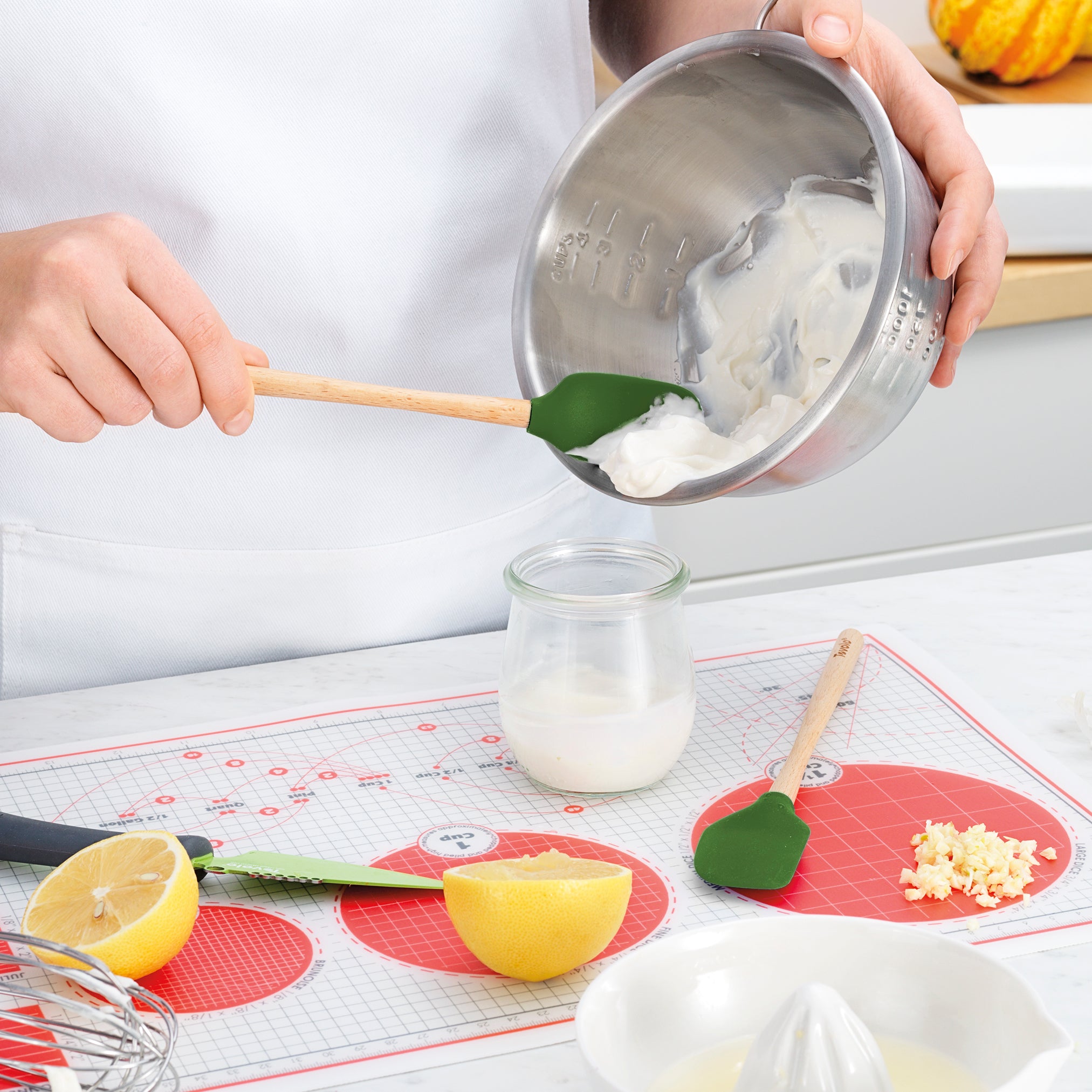 Using the green Flex-Core Mini Spat + Spoonula, a person spoons creamy yogurt from a metal bowl into a glass jar. A sliced lemon, knife, citrus juicer, chopped garlic, and another spatula rest on a red mat nearby.