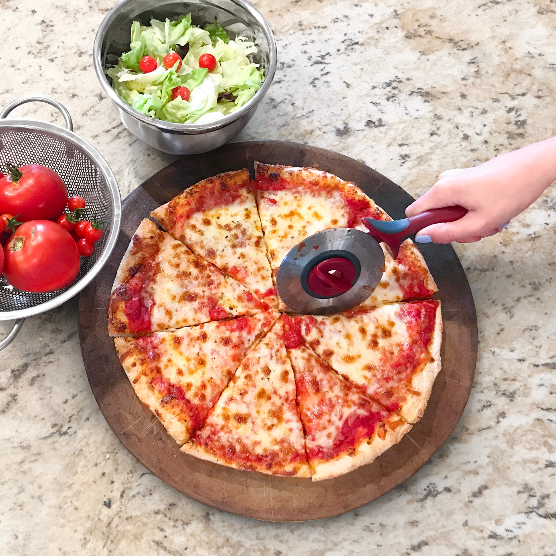 A hand uses a pizza cutter to slice a cheese pizza on a wooden board. Next to it, a Mixing Bowl holds salad with cherry tomatoes, while a colander contains fresh tomatoes on the light countertop.