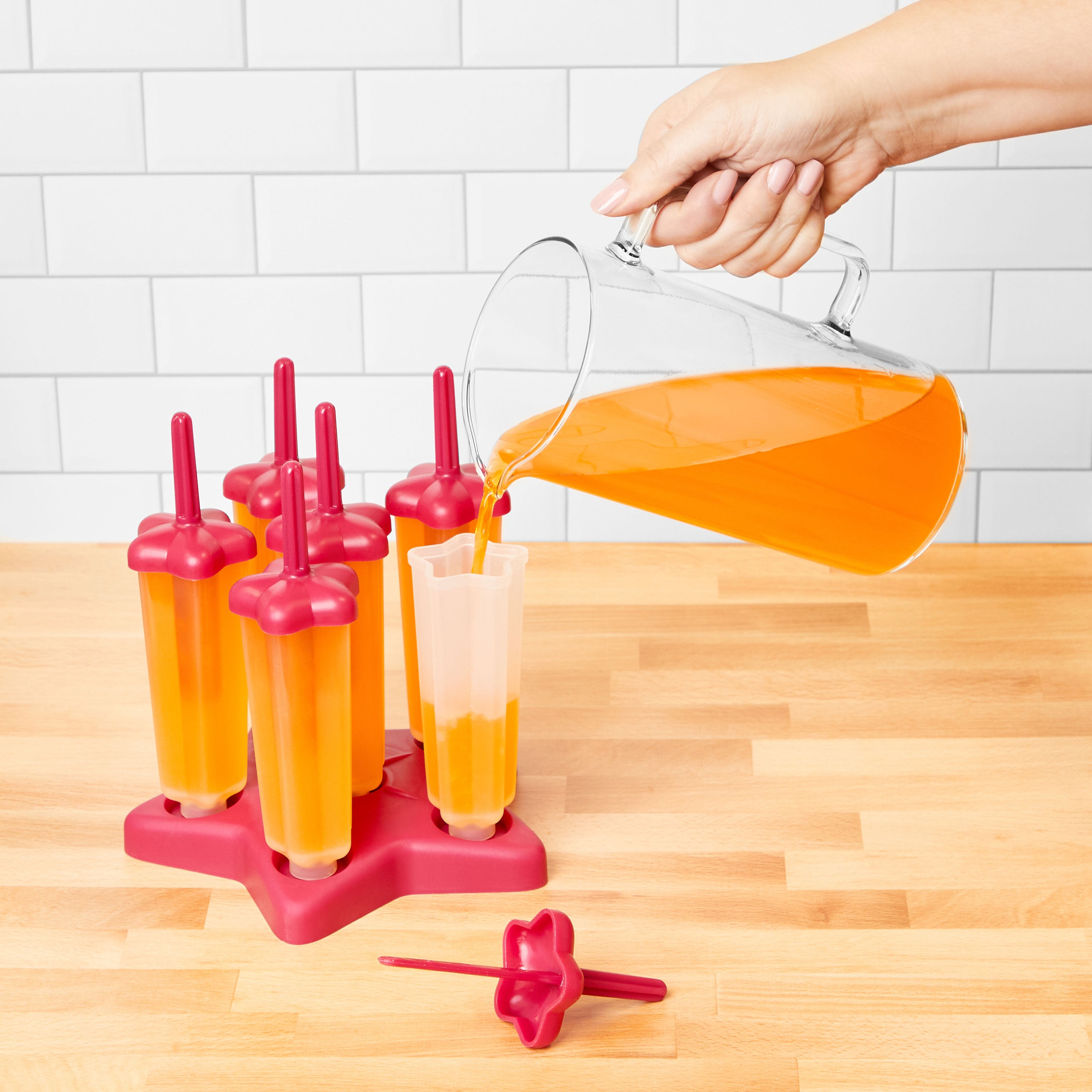 A hand pours orange juice from a glass pitcher into Pop Molds on a wooden countertop, with other juice-filled molds resting in a slimline tray and a white tile wall in the background for making homemade frozen treats.