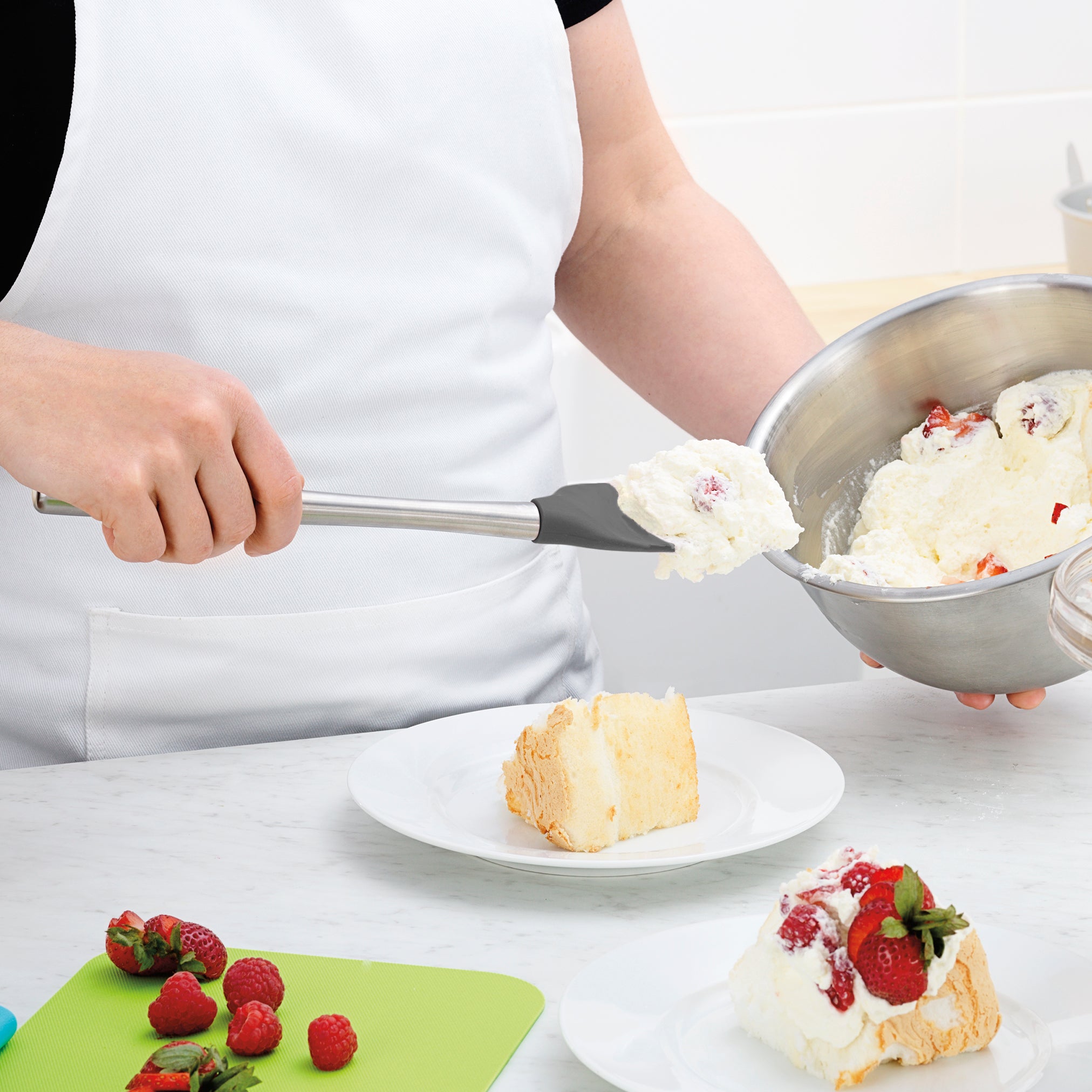 Wearing a white apron, a person uses the Flex-Core Spoonula to scoop whipped cream with strawberries from a metal bowl onto cake, while fresh strawberries and raspberries sit on a green cutting board nearby.