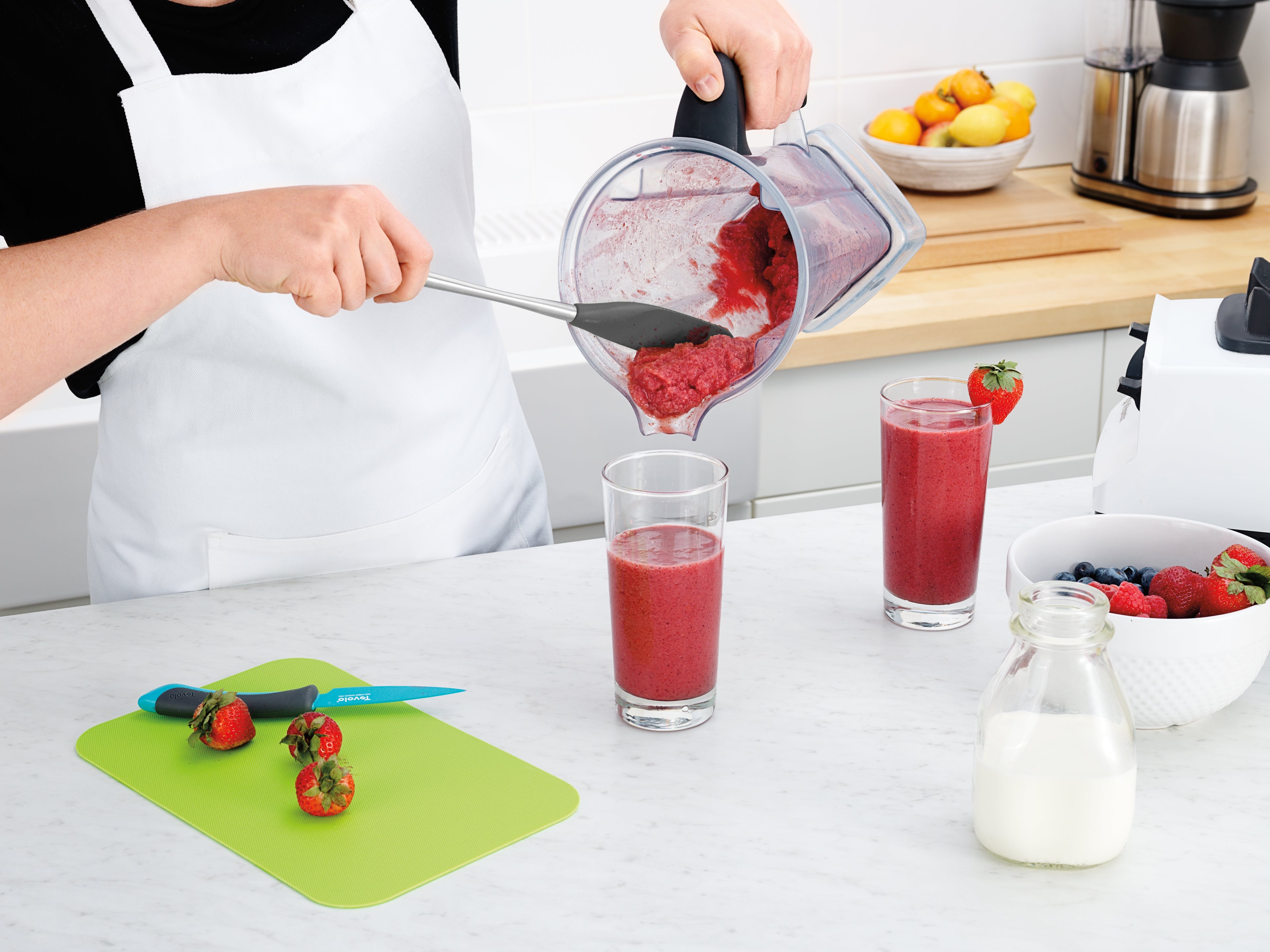 Wearing a white apron, a person uses the Flex-Core Jar Scraper to pour a thick red smoothie from a blender into a glass. On the white kitchen counter are two filled glasses, strawberries, a berry bowl, a knife, and a milk bottle.