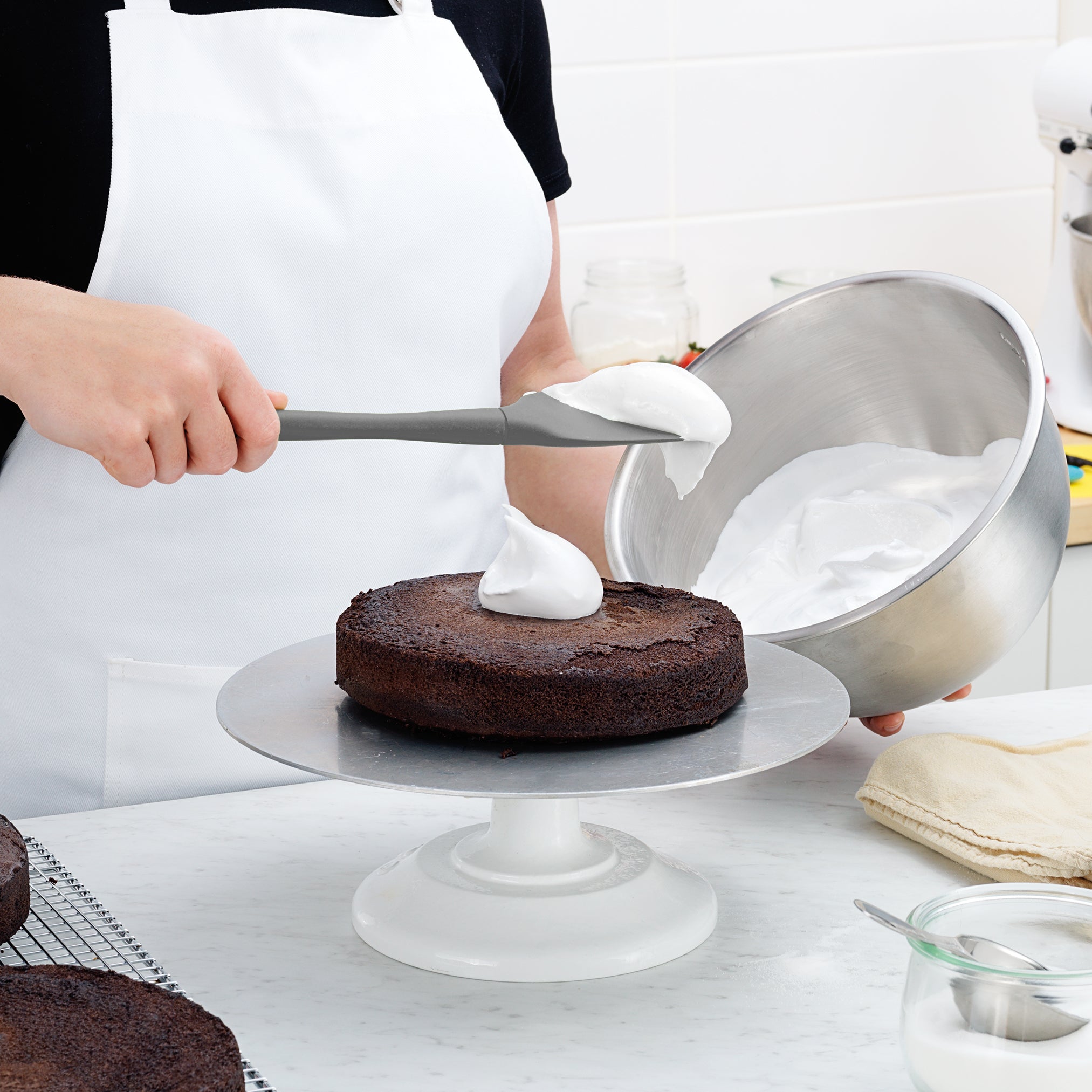 A person in a white apron uses the Flex-Core Spoonula to spread whipped white frosting onto a round chocolate cake, holding a metal mixing bowl with more frosting on hand.