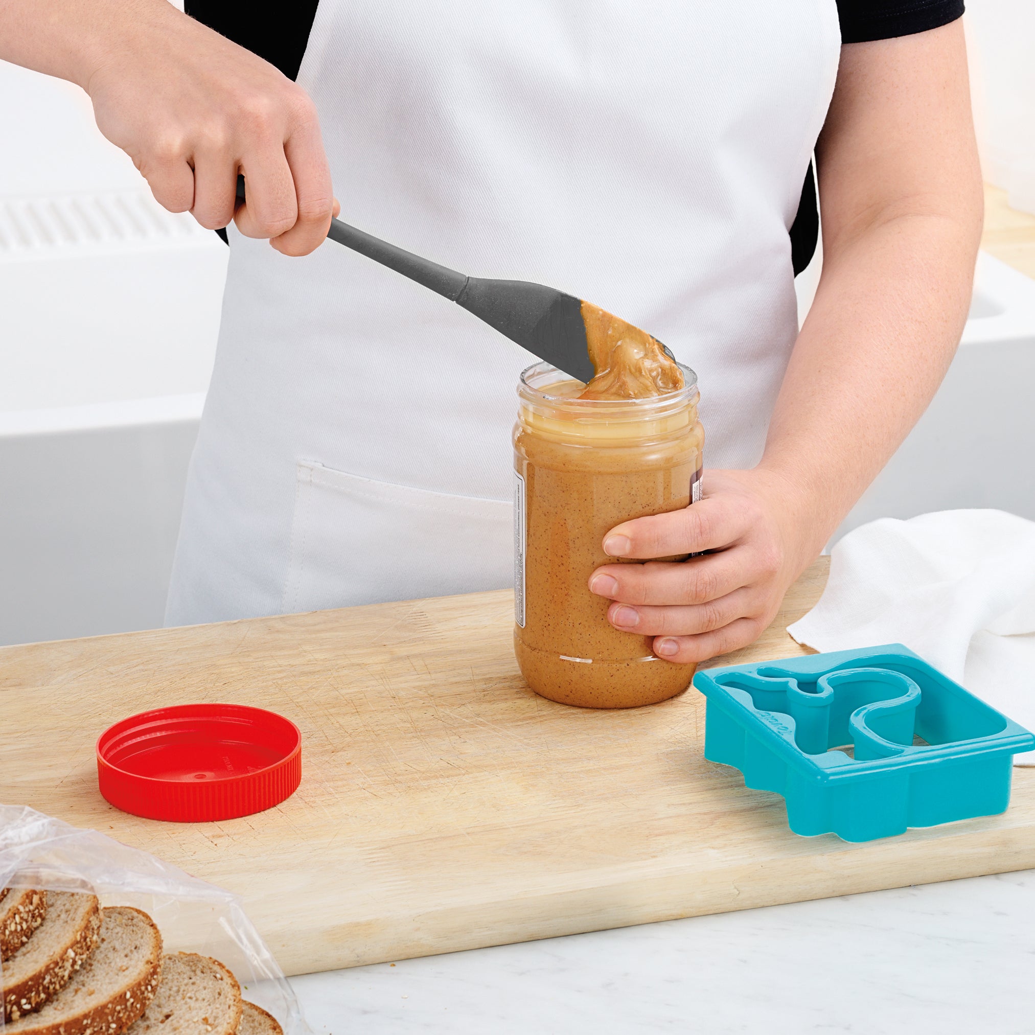 Wearing a white apron, someone uses the Flex-Core Jar Scraper to scoop peanut butter from a jar. A red lid, sliced bread, and a blue sandwich cutter rest on the wooden cutting board, ready for sandwiches.