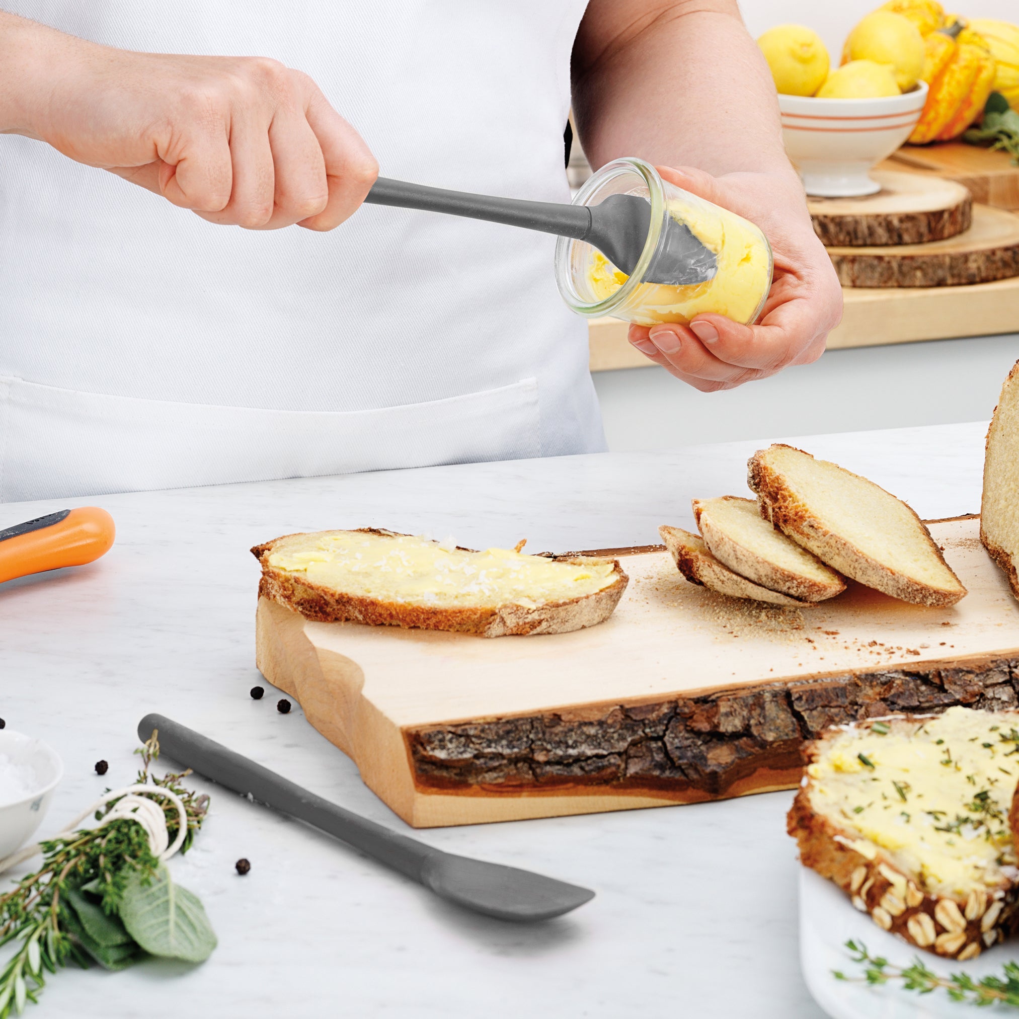 A person uses the Flex-Core Mini Spat + Spoonula to spread yellow butter on rustic bread slices atop a wooden cutting board. Fresh herbs, a butter knife, and other kitchen items sit on a white countertop.