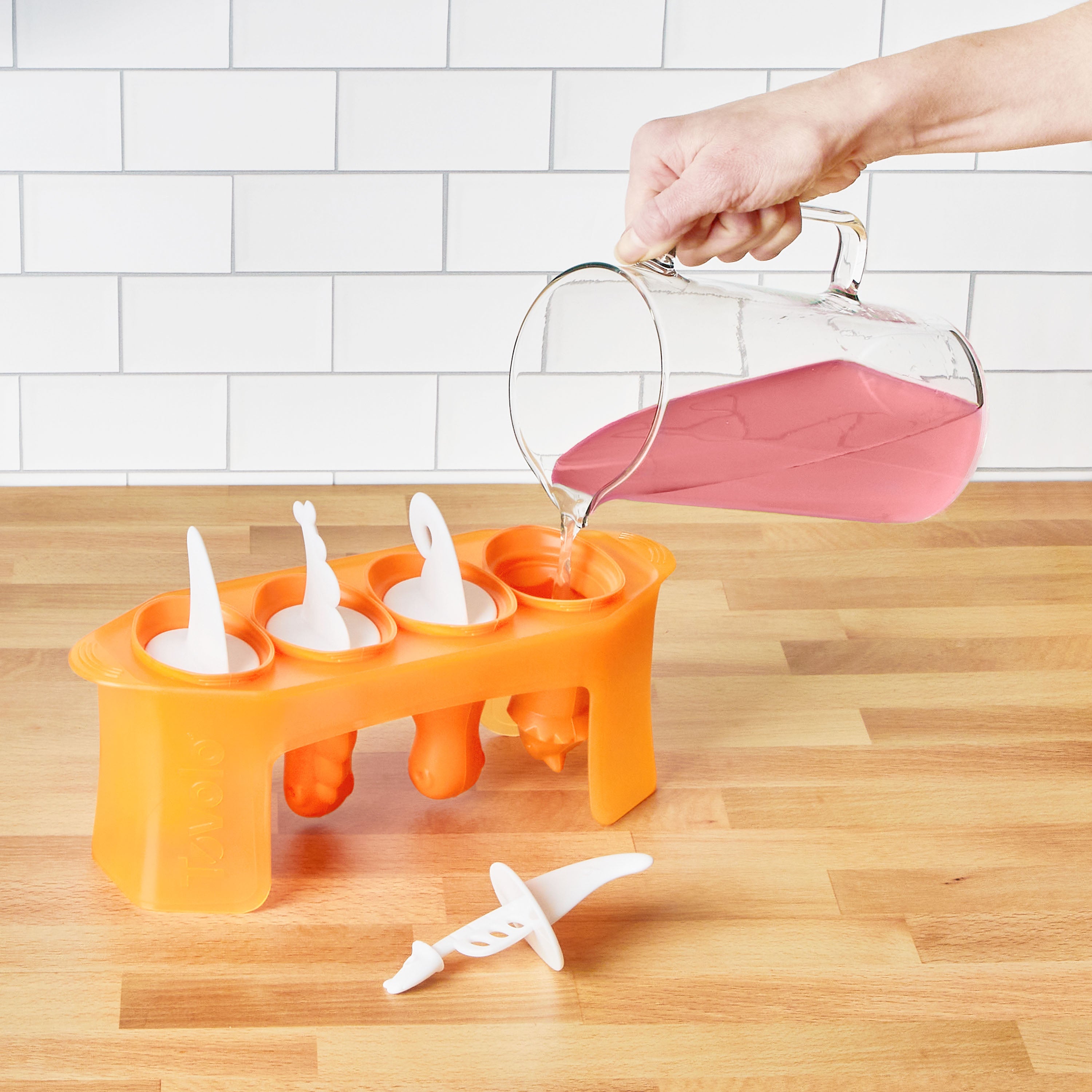 A hand pours pink liquid from a pitcher into an open Pop Molds orange popsicle mold on a wooden countertop; three Pop Molds are closed, one is open, all set against a kitchen with a white tile backsplash.