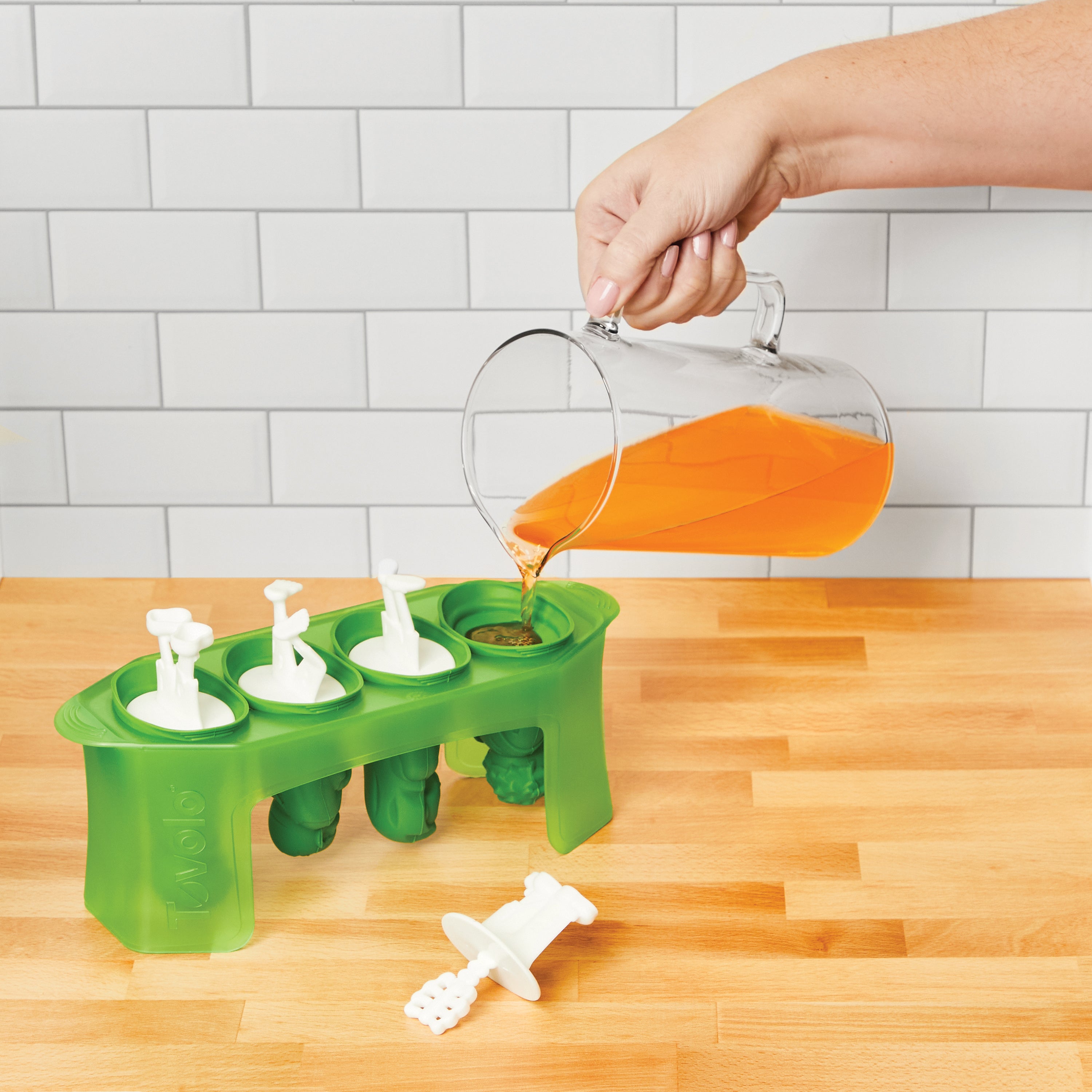 Person pouring liquid into a green silicone mold on a wooden surface with a tiled wall background.